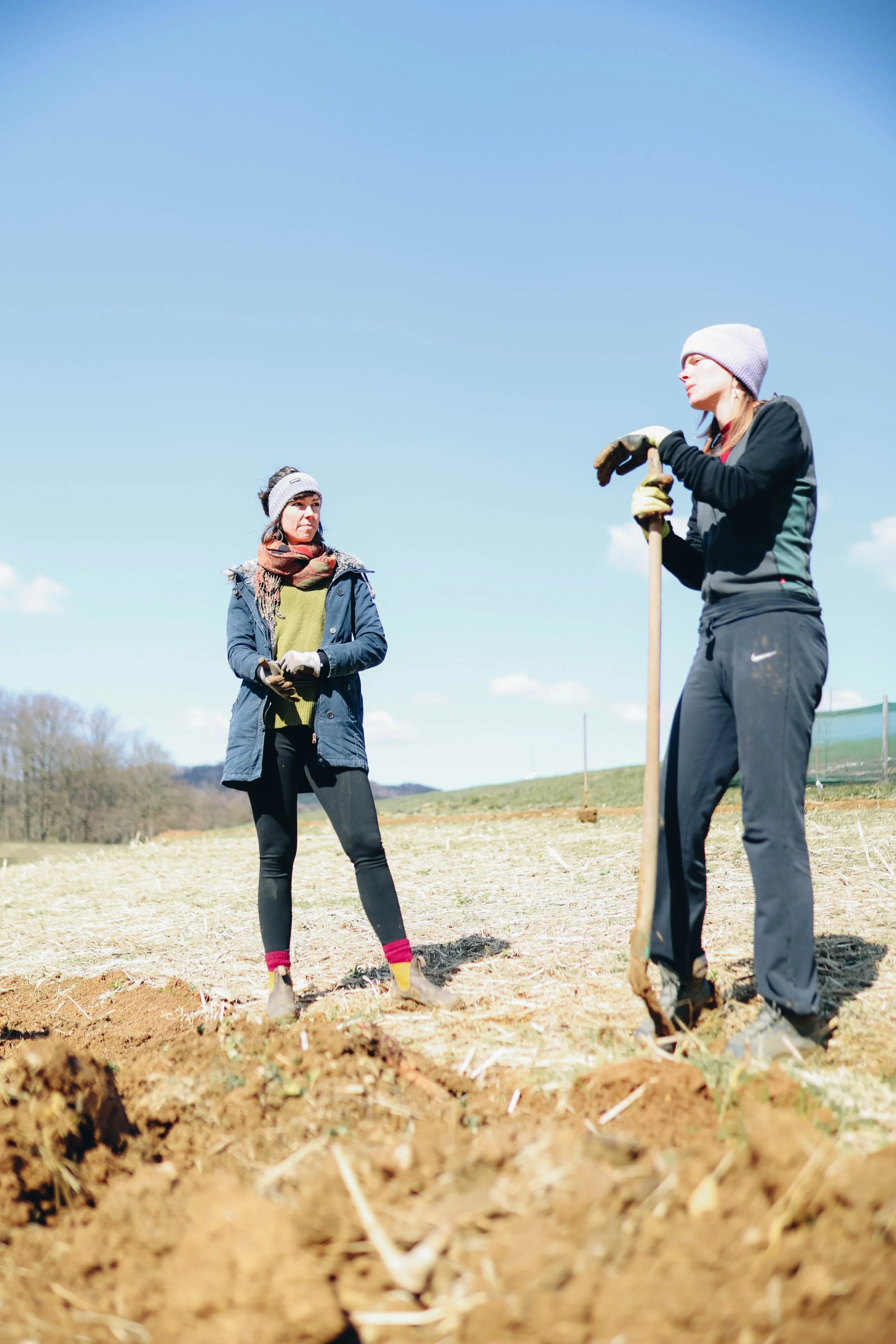 Two women in outdoor clothing and hats working together outdoors on a sunny day, with one holding a shovel.
