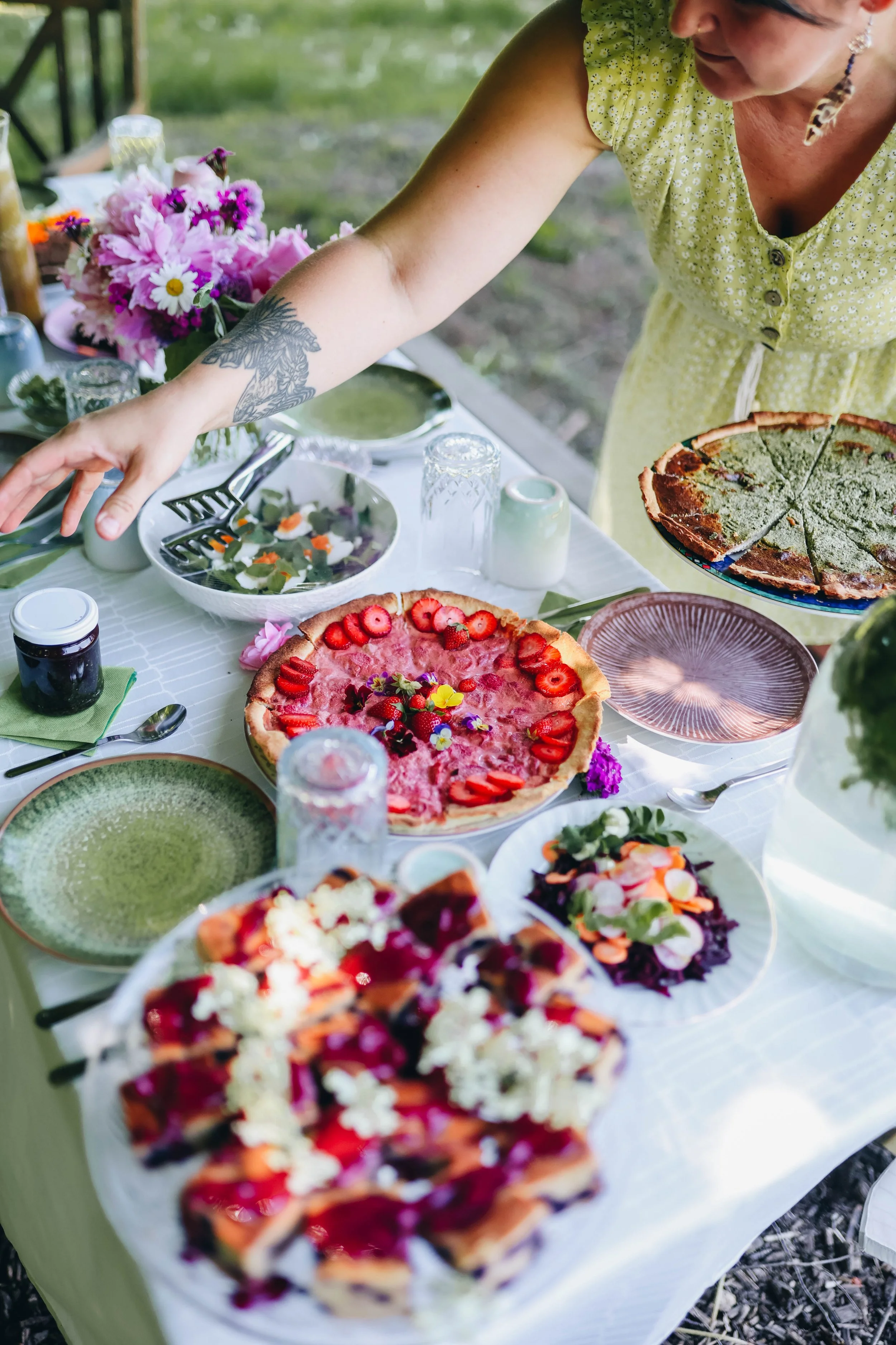 Table set outdoors with assorted desserts including berry-topped tarts and fruit salads, as a woman in a yellow dress reaches for food.