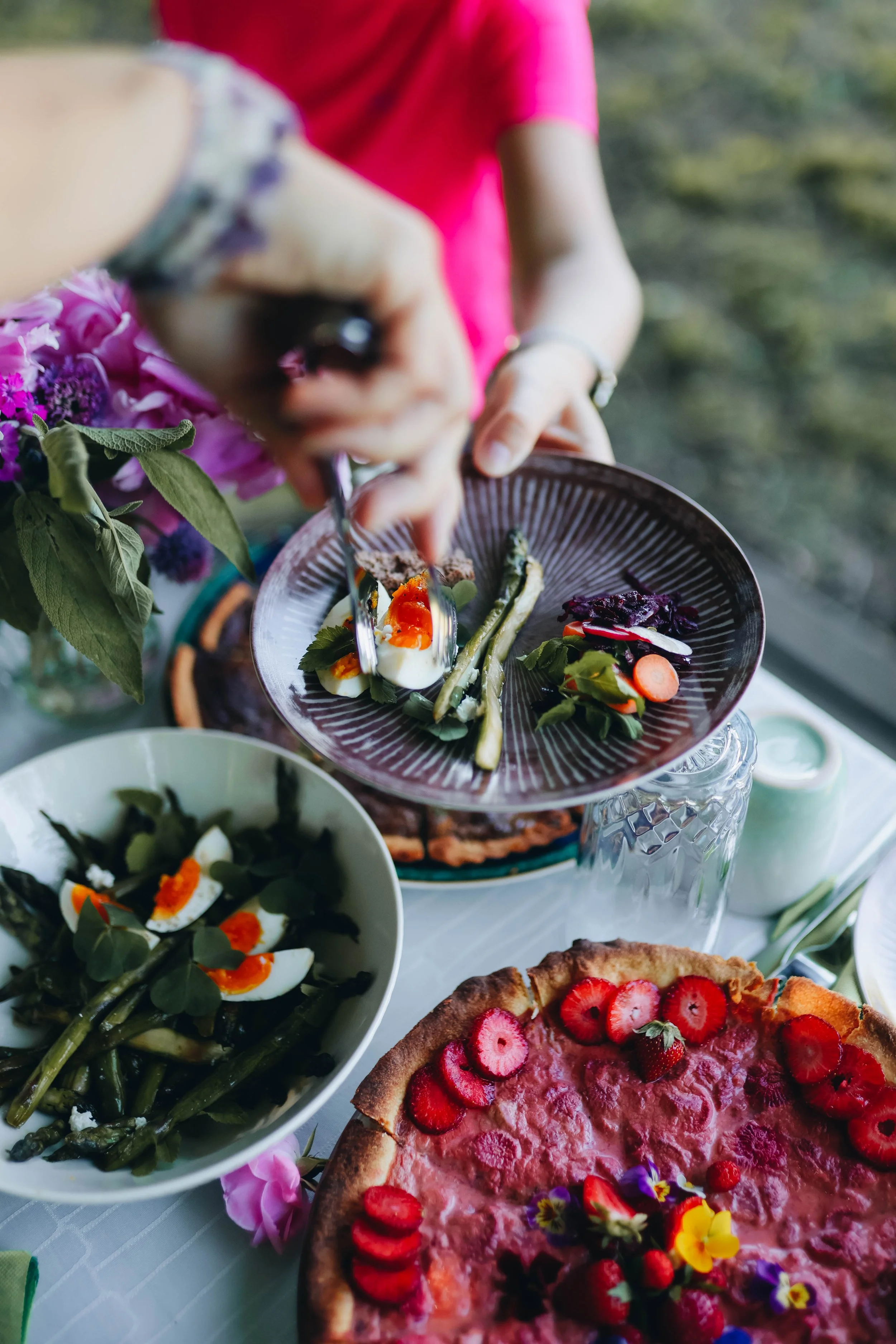 Person serving various salads and a strawberry-topped dessert at a table with flowers.