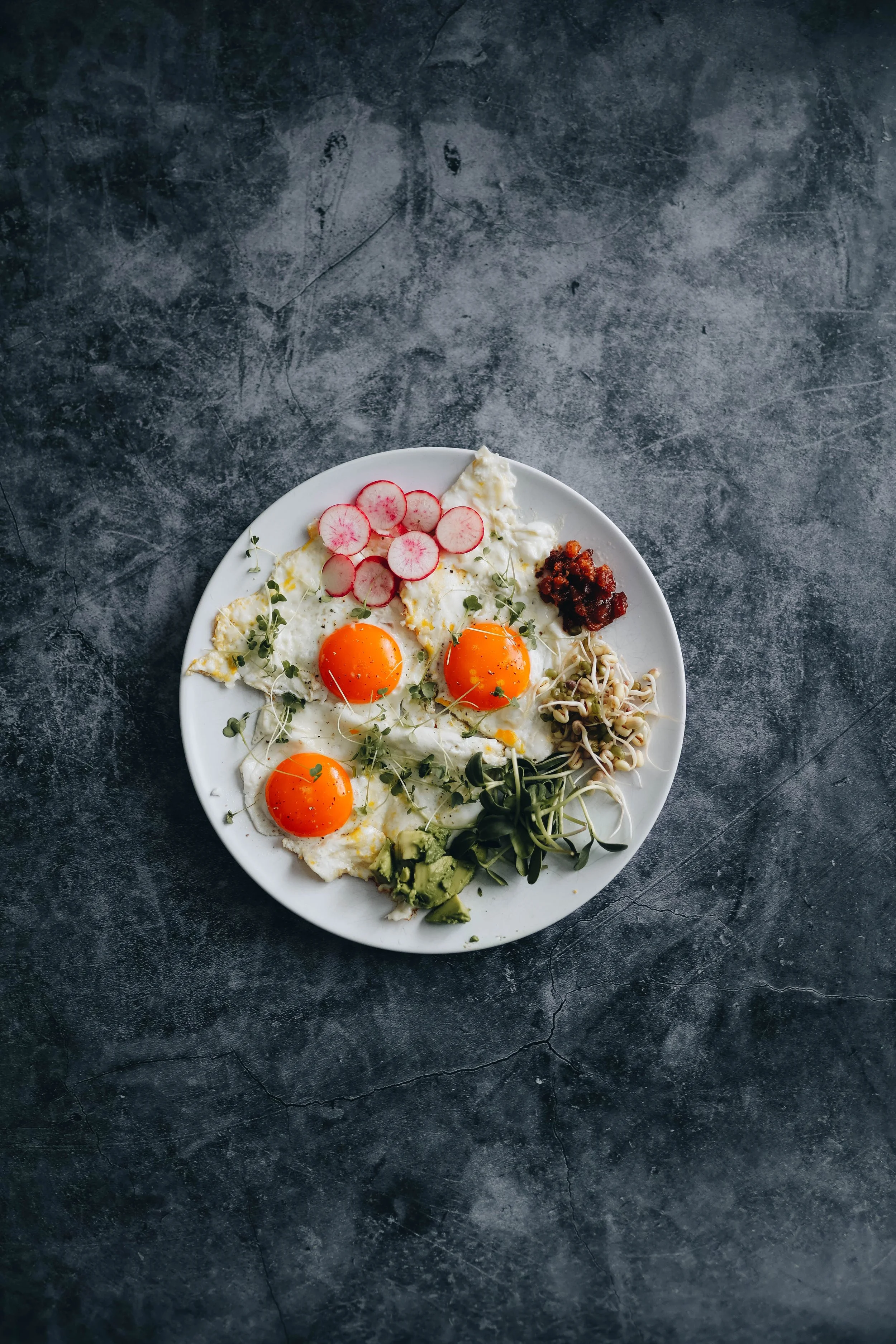 A white plate with sunny side up eggs, radish slices, sprouts, diced avocado, and a small serving of jam, set on a textured dark gray surface.