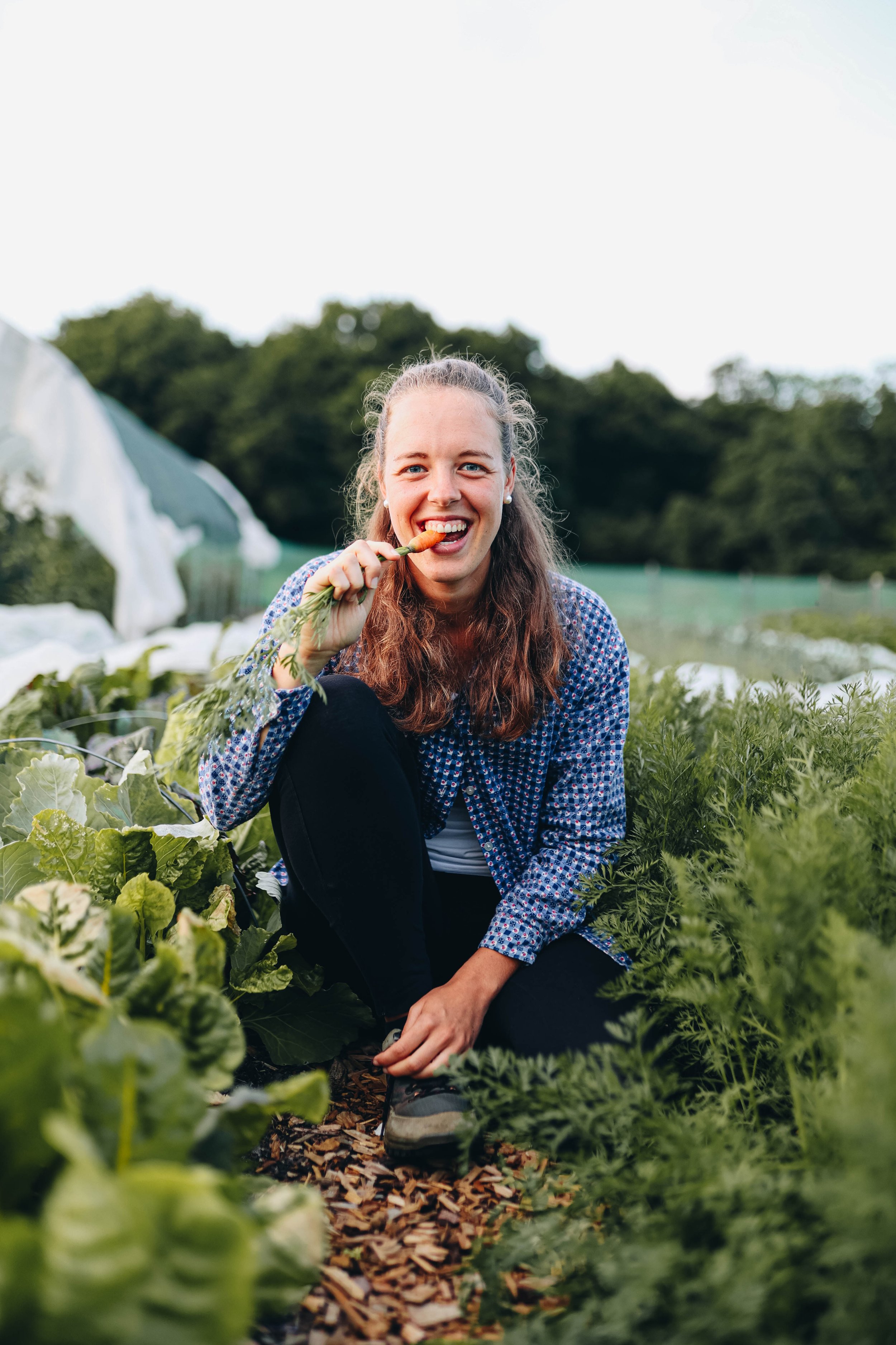 A woman smiling and eating a carrot while crouching in a vegetable garden surrounded by green plants.