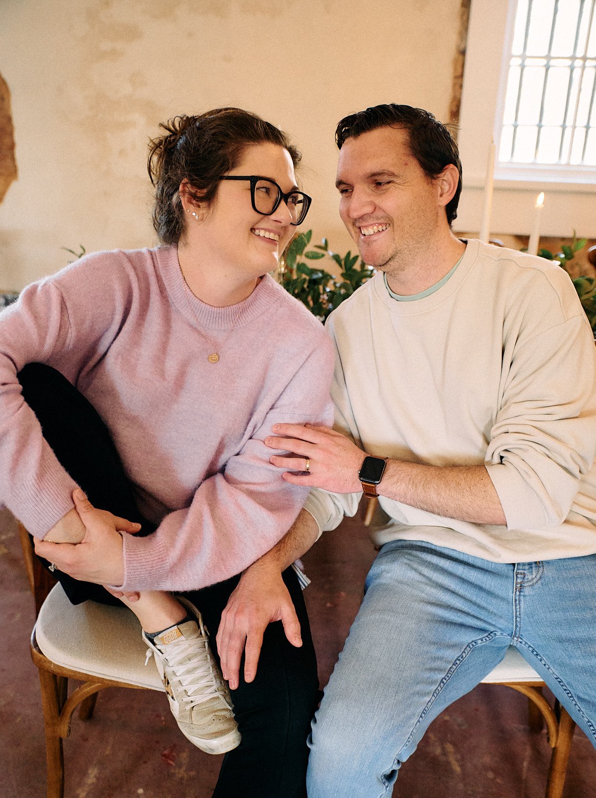 A woman with glasses and a man are sitting close together, smiling and touching each other's arms. The woman is wearing a pink sweater and the man is wearing a cream-colored sweatshirt and jeans.