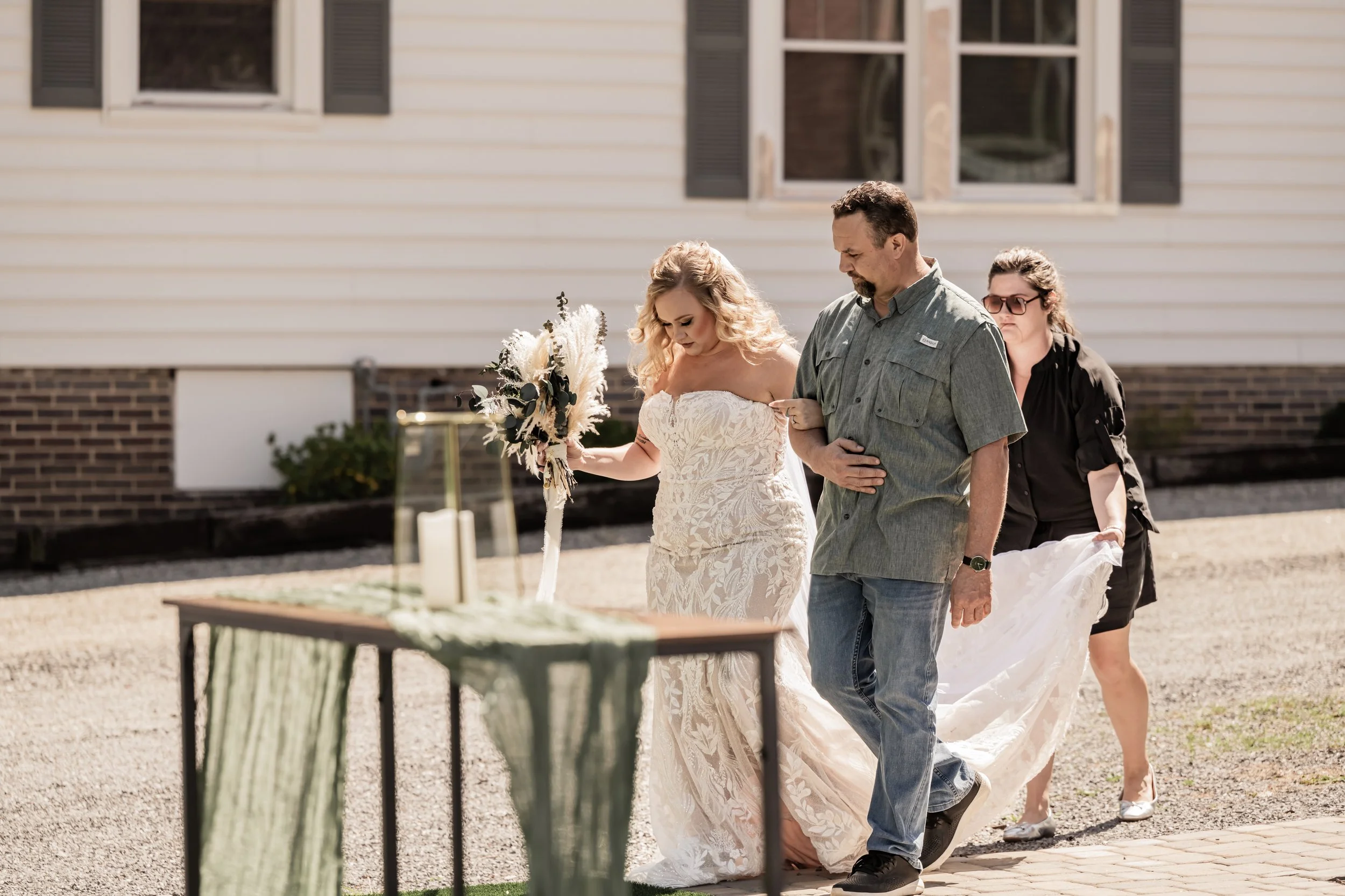 A bride in a white lace wedding dress holding a bouquet and walking outdoors with the support of a man in a green shirt and jeans, while a woman in black adjusts her dress behind them.