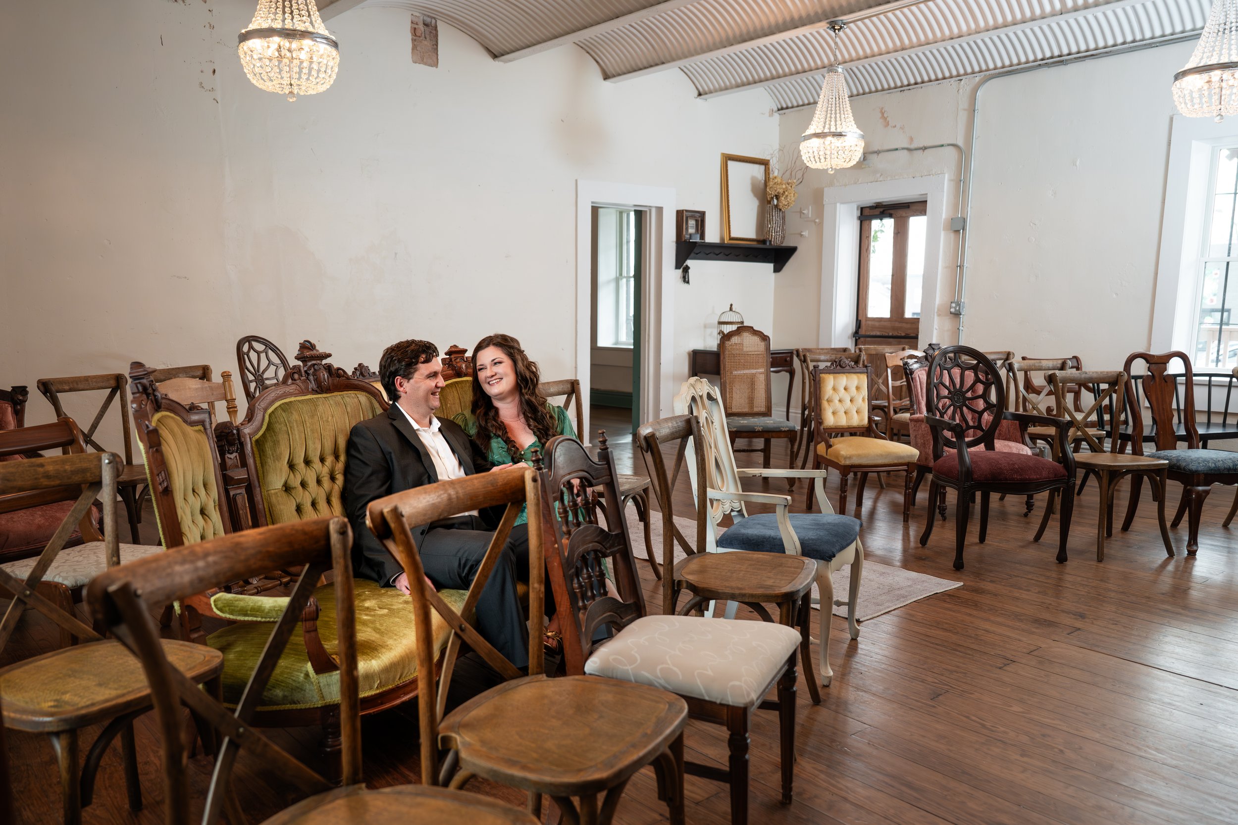 A man and woman sitting on a vintage green velvet sofa, smiling at each other, in a room filled with various wooden chairs and furniture, with chandeliers hanging from the ceiling.