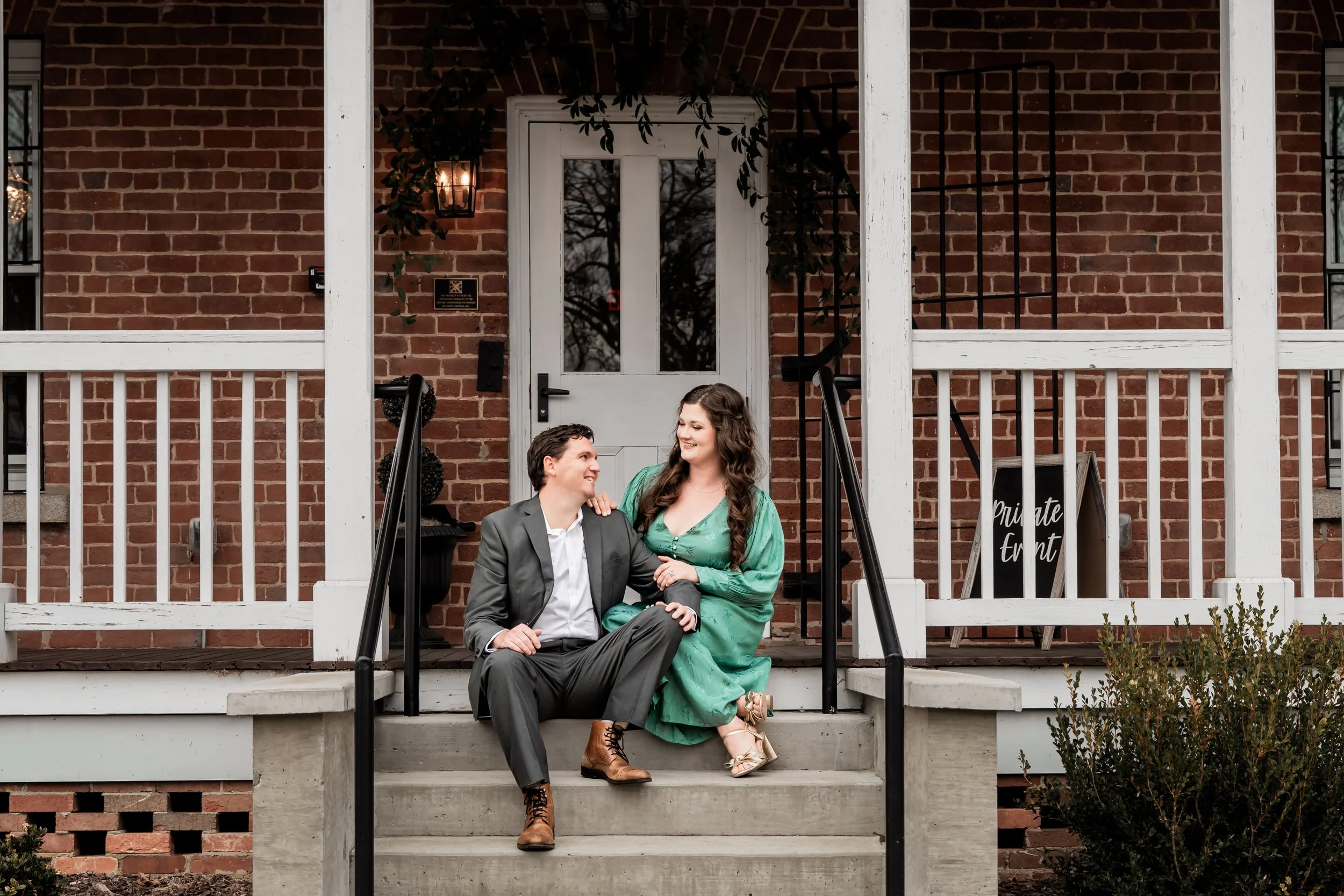 A man and woman sitting on the front steps of a brick house, smiling and looking at each other. The man wears a gray suit and brown boots, and the woman wears a green dress and beige heels. The house has a white door, black railings, and a sign that 