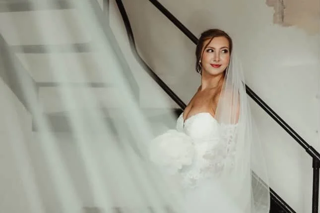 Bride in a white wedding dress with a veil standing on a staircase, looking to the side with a smile at The Historic Dallas Jail.