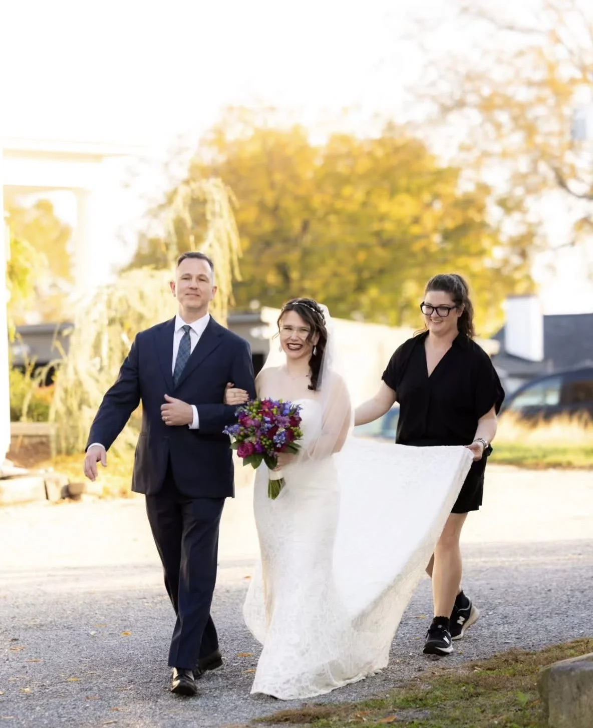 A bride holding a bouquet of flowers walking with a man in a suit, and a woman assisting with her gown outdoors with autumn trees in the background.