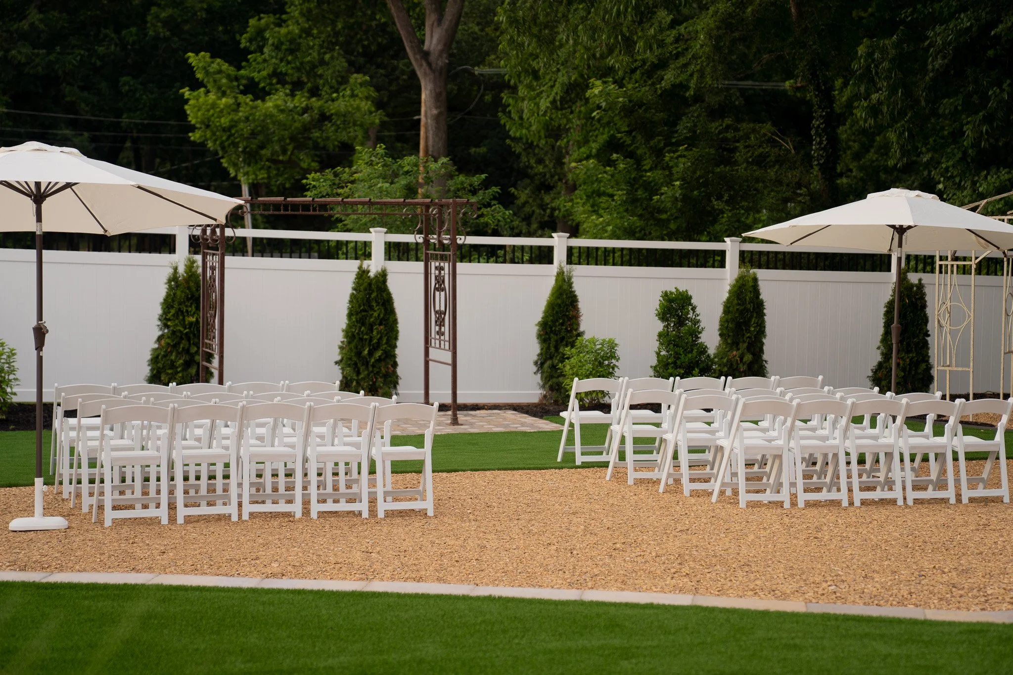 The Historic Dallas Jail Outdoor wedding or event setup with white chairs arranged on a gravel ground, white umbrellas, green shrubs, and a white fence with a gate in the background, surrounded by tall green trees. Charlotte Historic Wedding Venue
