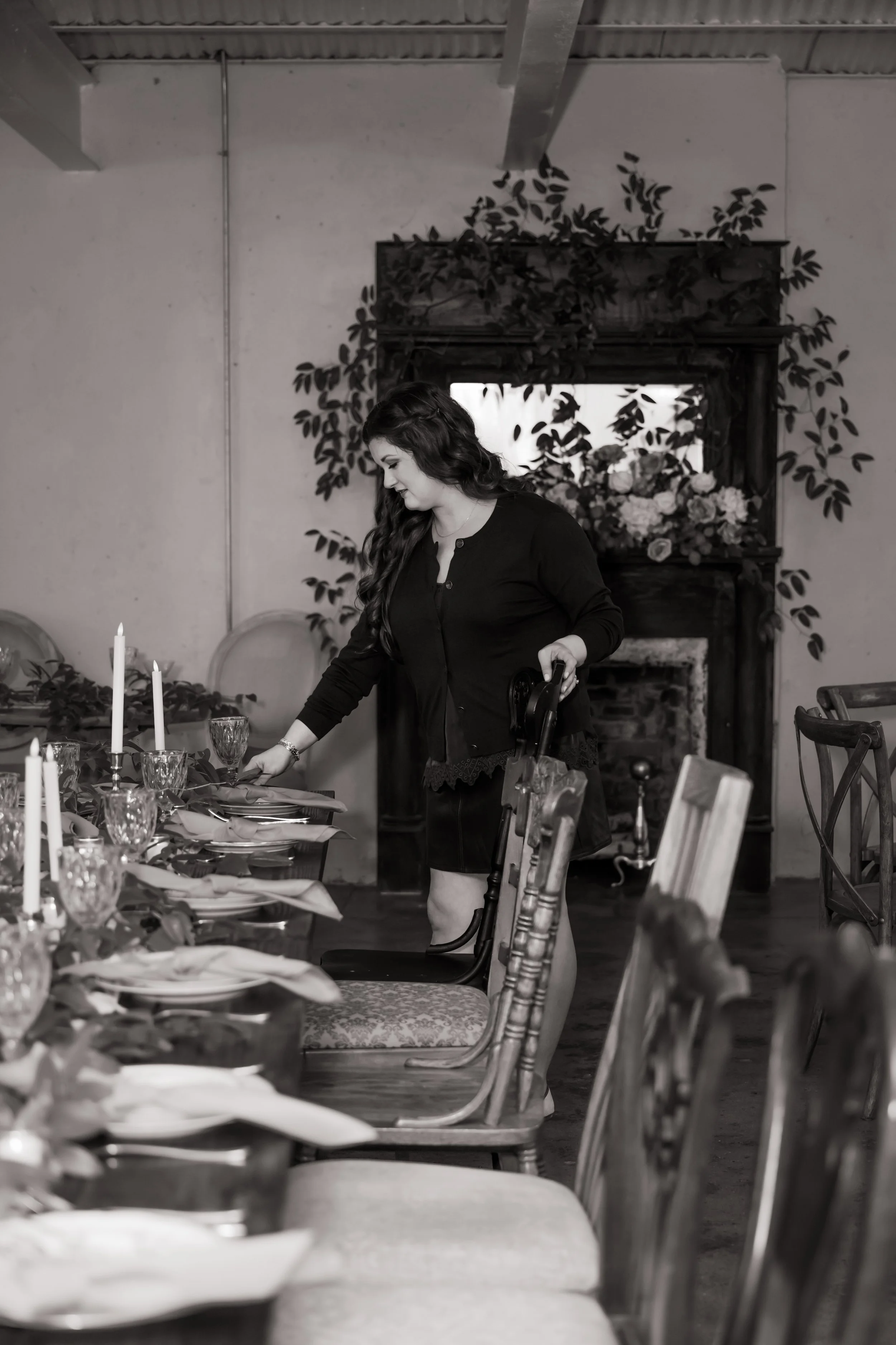 Woman in black dress setting a dining table in a decorated room, black and white photo.