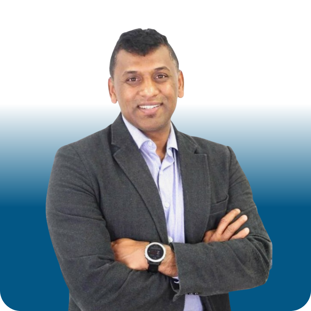 A man with short black hair, wearing a dark gray blazer, light blue shirt, and a wristwatch, standing with arms crossed and smiling at the camera against a plain white background.