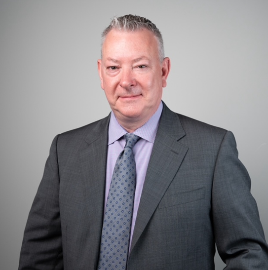 Professional headshot of a middle-aged man wearing a grey suit, light purple shirt, and patterned tie, standing against a neutral grey background and looking directly at the camera.