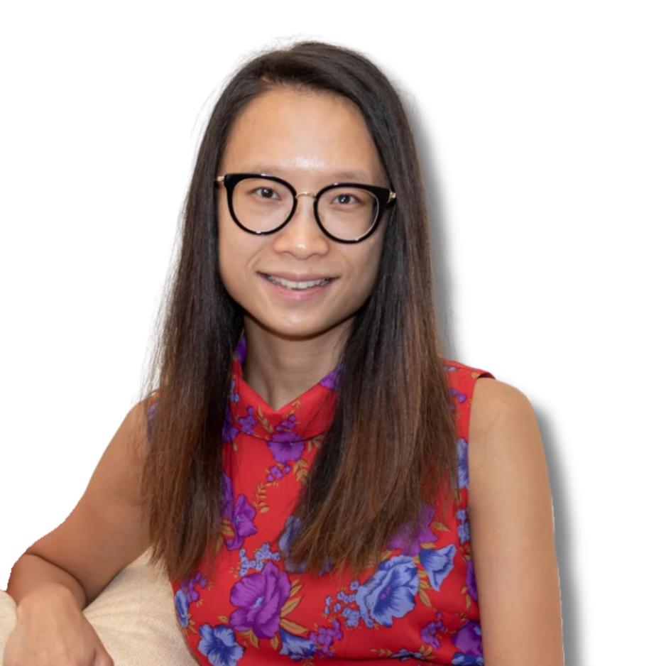 A woman with long dark hair, wearing black-rimmed glasses, smiling, dressed in a red floral dress, sitting on a beige cushion on a white couch.