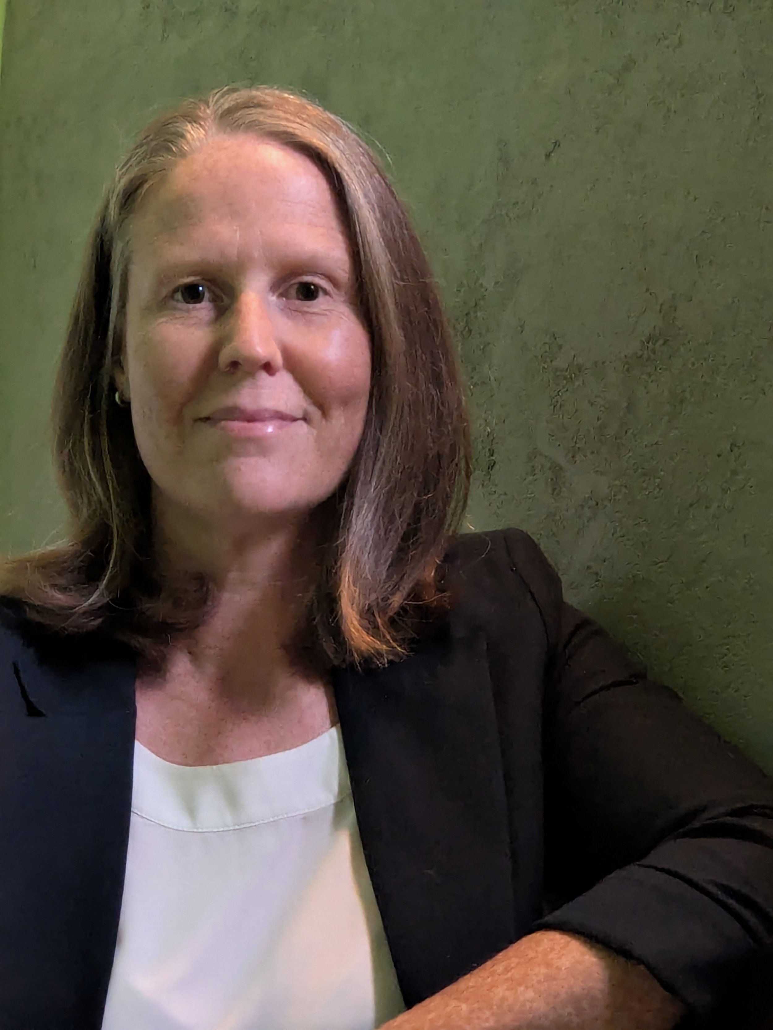 A woman with long brown hair, wearing a black blazer and white top, smiling softly, against a green textured background.