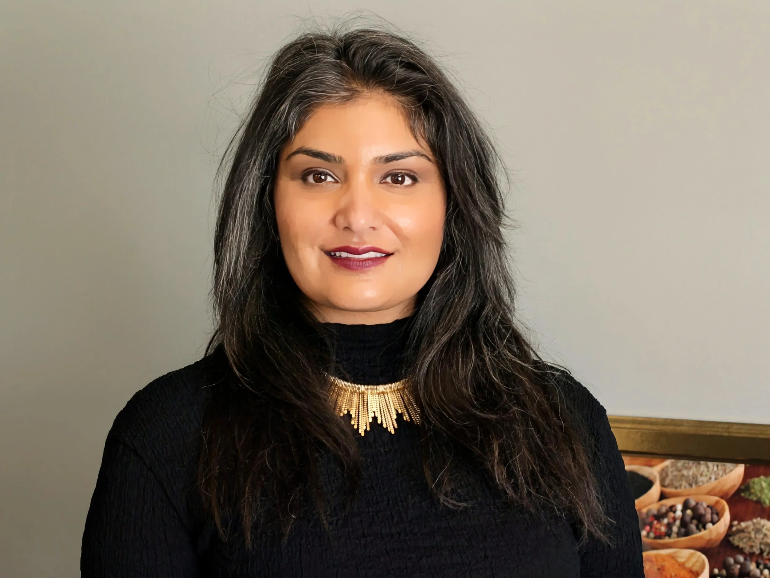 A woman with dark hair, a black turtle neck with a gold necklace and dark rouge lips