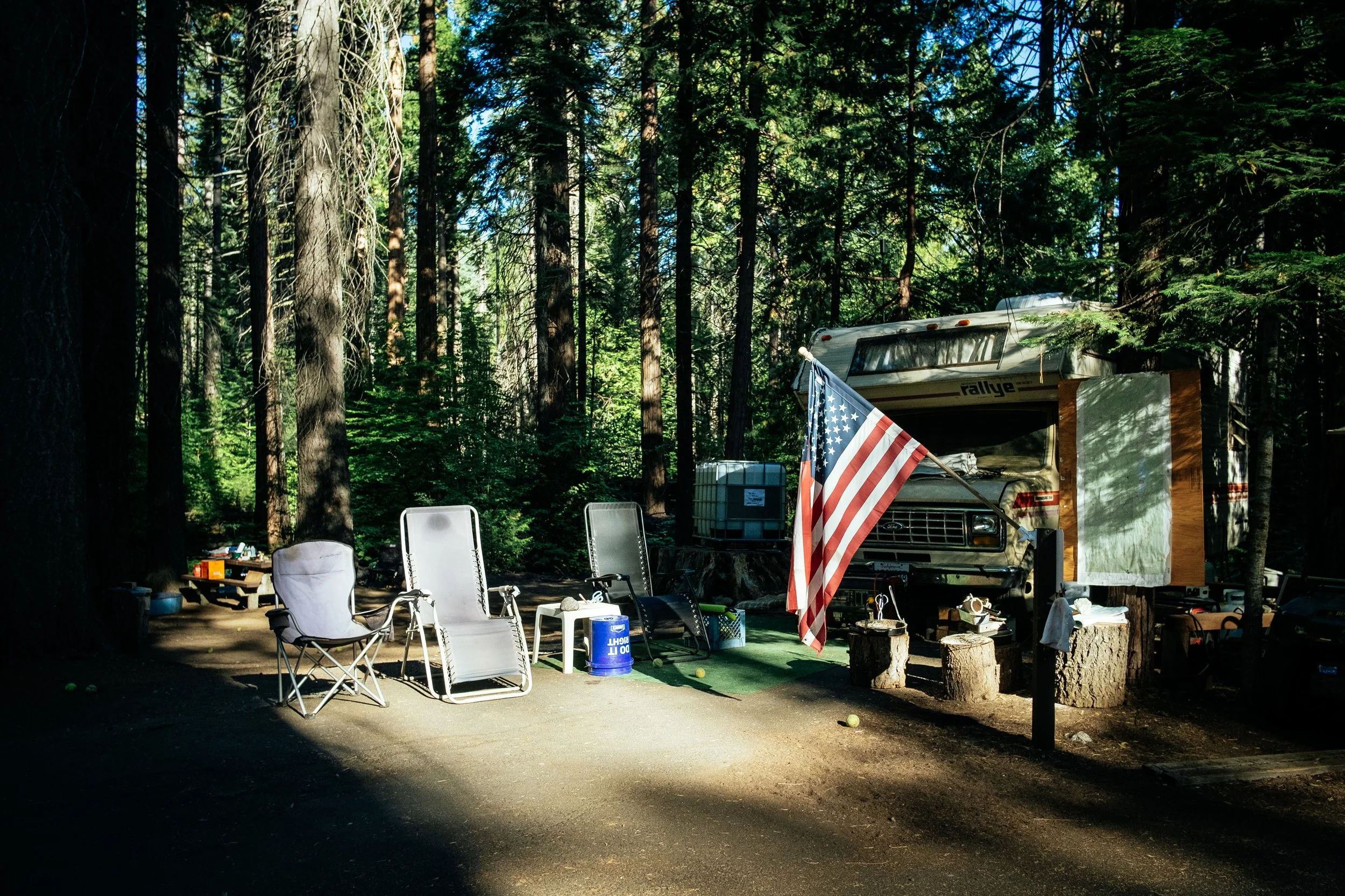 Campsite with trailer and folding chairs among tall trees, from Andrea Marzagalli's photography series
