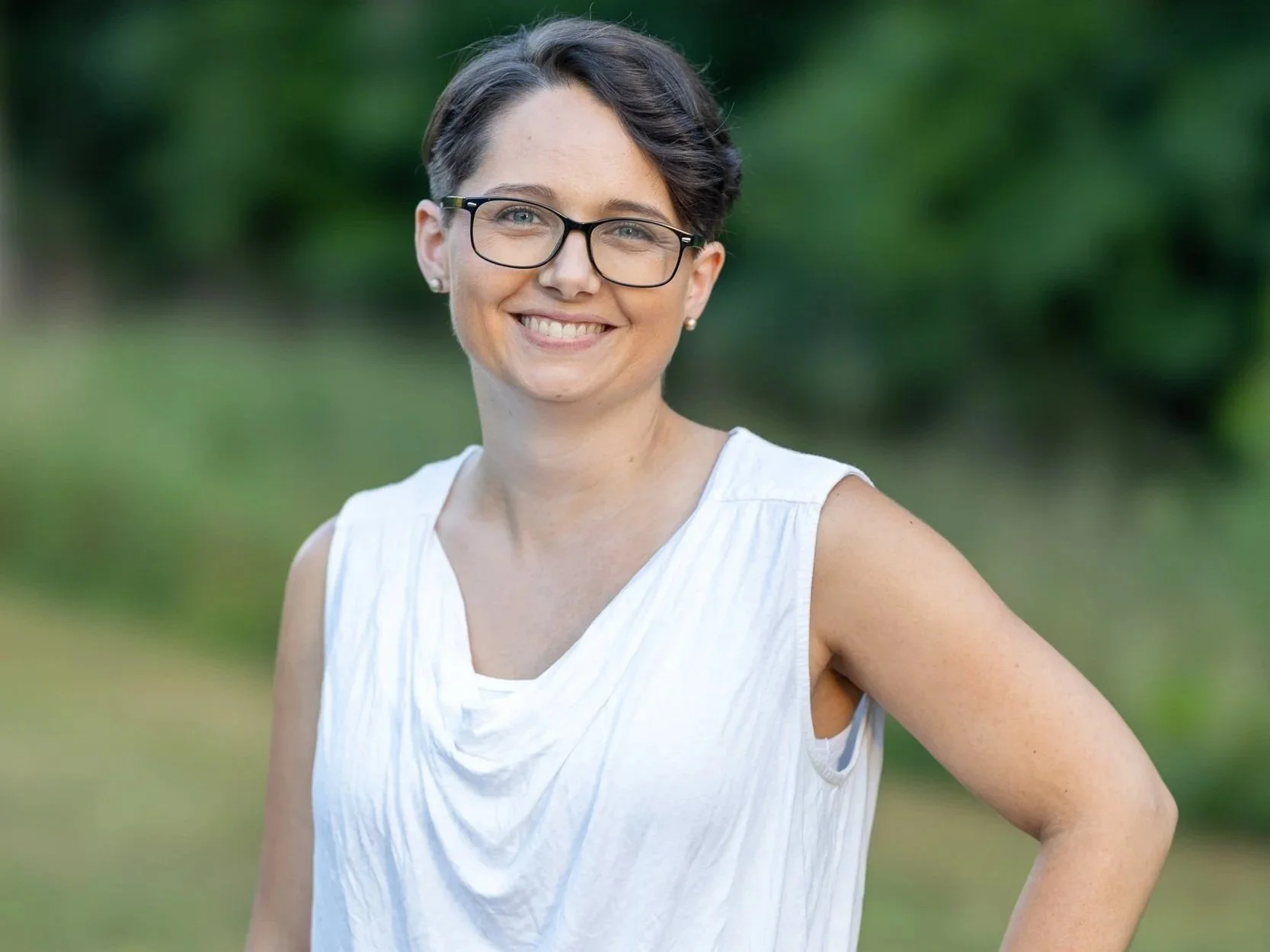 Tara White, is  a Birth Doula serving Raleigh, NC and the surrounding area. In this photo she appears as a woman with short dark hair, glasses, and pearl earrings smiling outdoors with a green, blurred background.