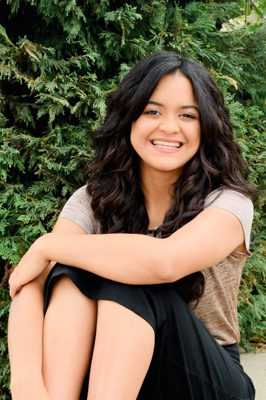 A young woman with long dark wavy hair smiling, sitting with arms around her knees, in front of green shrubbery.