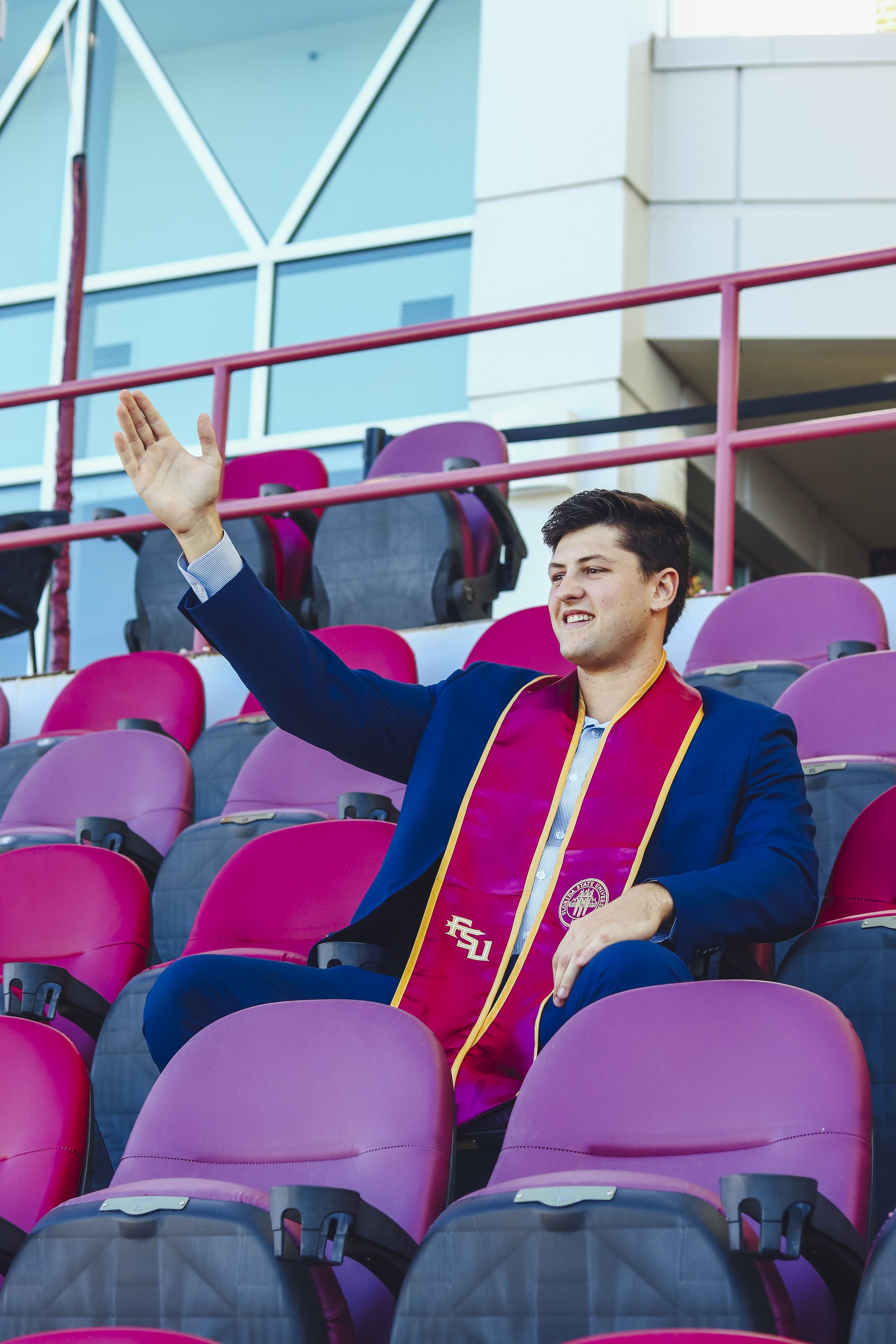 Smiling woman in graduation attire with a maroon stole, standing on a pathway surrounded by greenery.