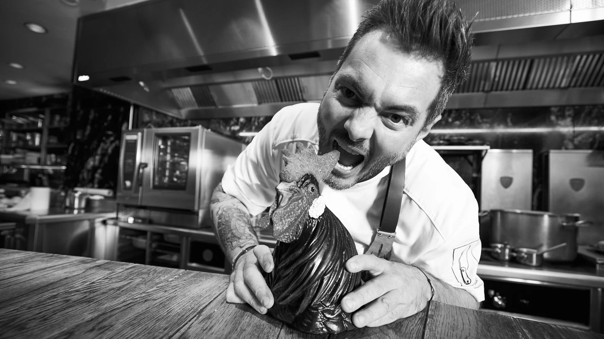 Un cocinero con barba y tatuajes, sosteniendo una figura de cerdo de chocolate y haciendo una expresión divertida en una cocina moderna.