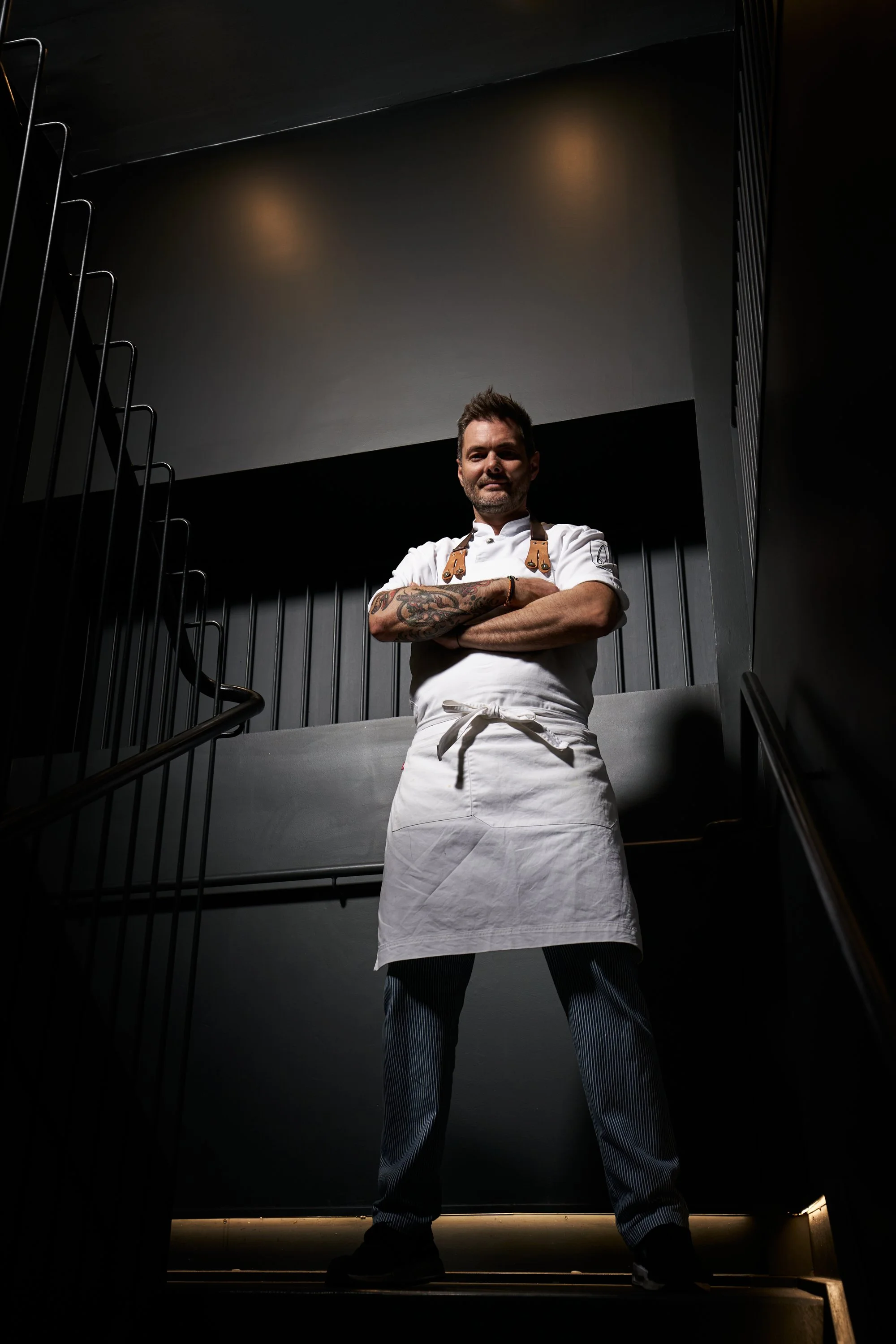 Chef Jose del Hotel Gastronómico Eunice con brazos cruzados en un ambiente oscuro, vistiendo uniforme blanco y delantal, en la parte superior de unas escaleras.