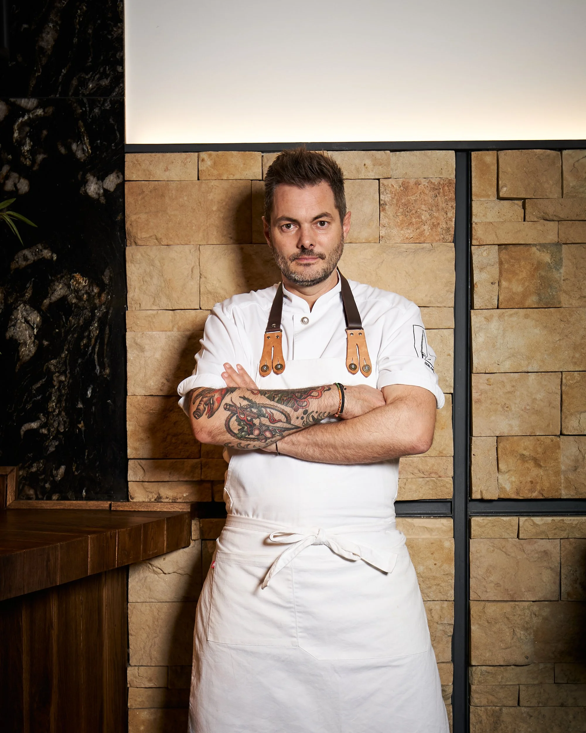 Chef masculino en uniforme blanco y delantal, con brazos cruzados, de pelo castaño oscuro y barba, posando en cocina con paredes de piedra beige y rincón con superficie de madera.