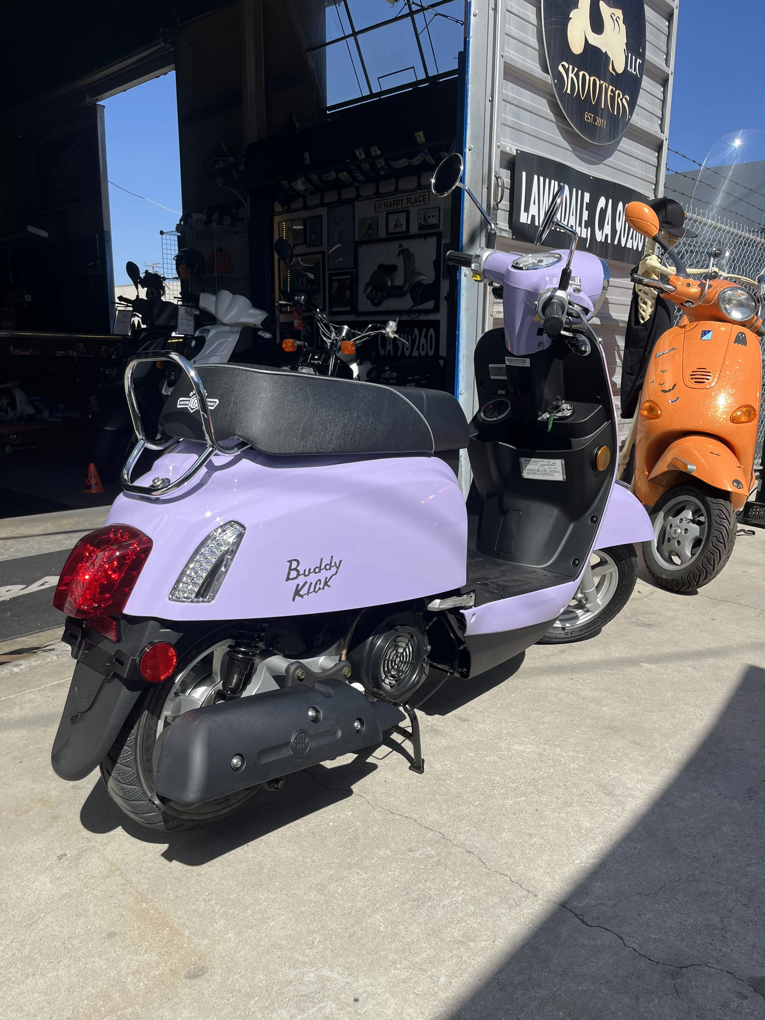 A small, purple electric scooter with a black seat and the words 'Buddy Kick' written on its side, parked outside a scooter shop.
