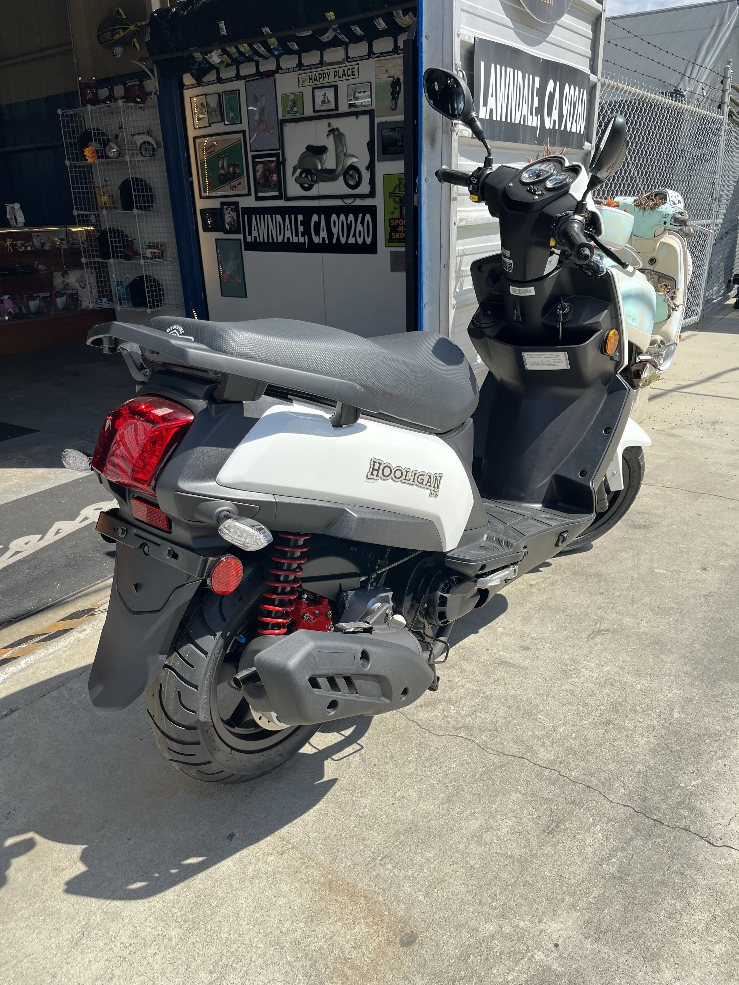 A Honda Scootier motorcycle with a white and gray body, black seat, and red suspension springs parked on a concrete sidewalk outside a small storefront with framed pictures and signs, including a sign for Lawndale, California.