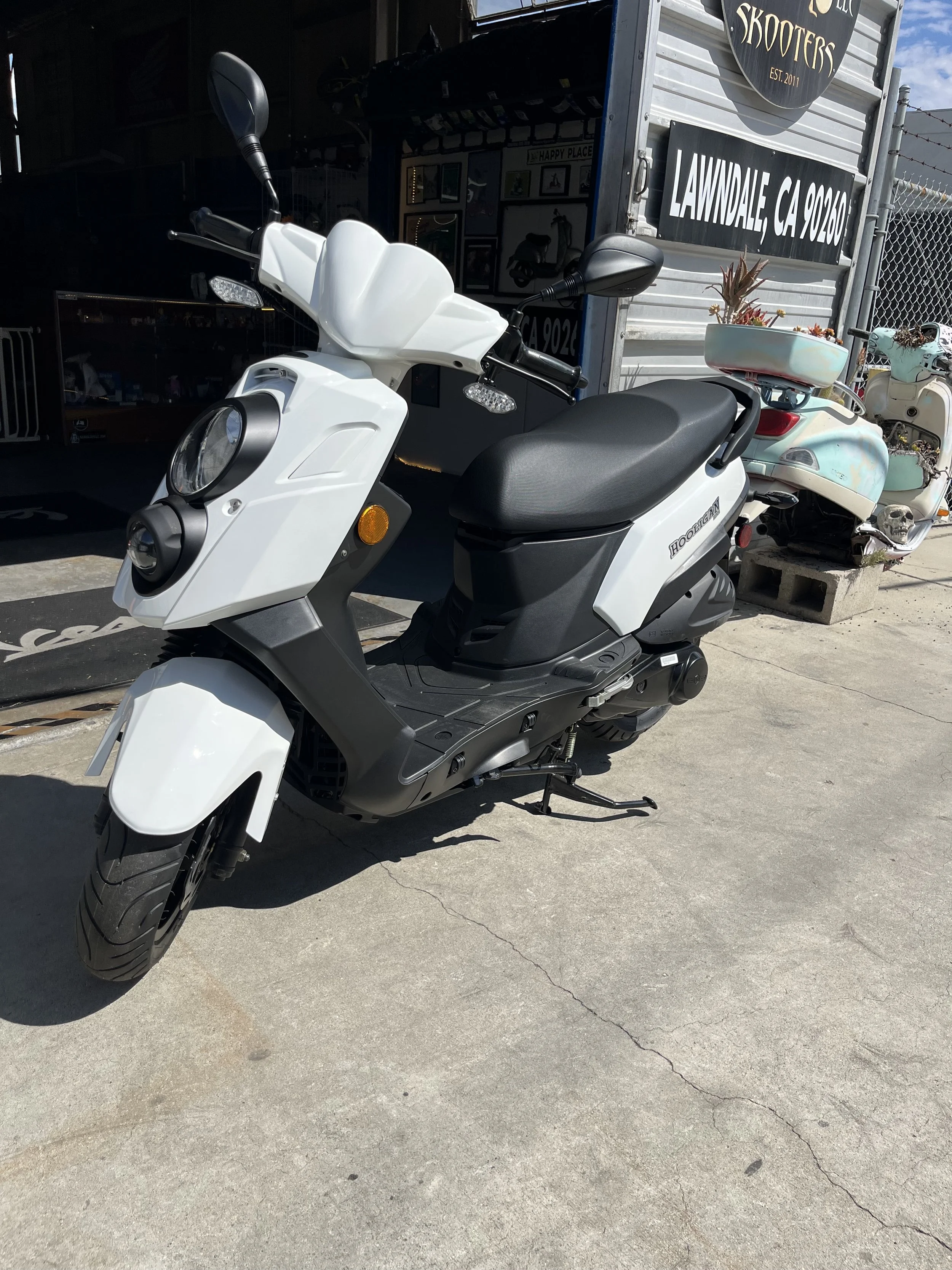 White electric scooter parked on a concrete sidewalk outside a shop with signs for skateboards and a store in Lawndale, California.