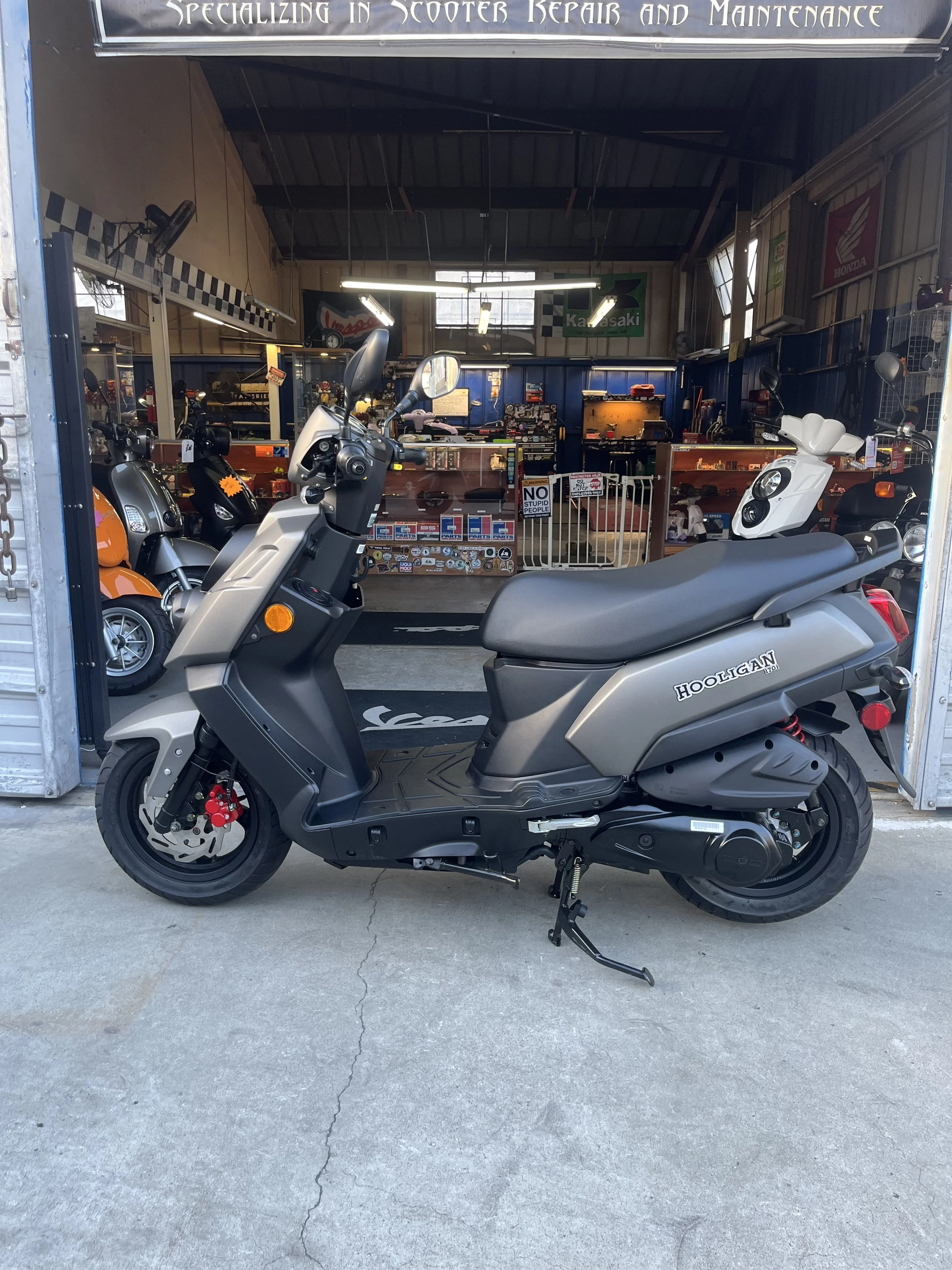 A gray Harley Quinn scooter parked outside a scooter repair shop, with various scooters and motorcycle parts inside the shop visible in the background.