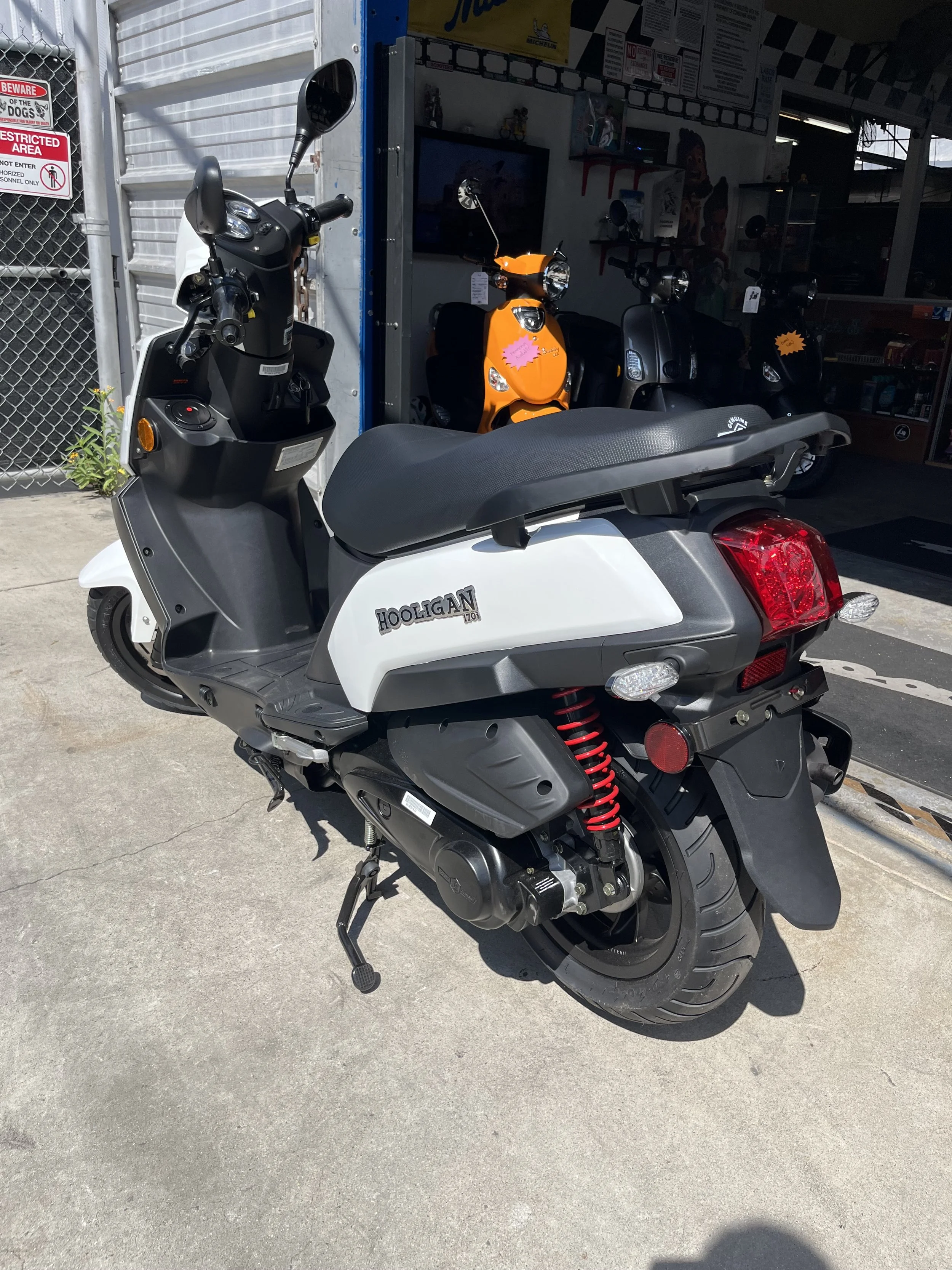 A white scooter with the label 'Hooligan 170' parked outside a shop, with over a row of other scooters inside the shop.