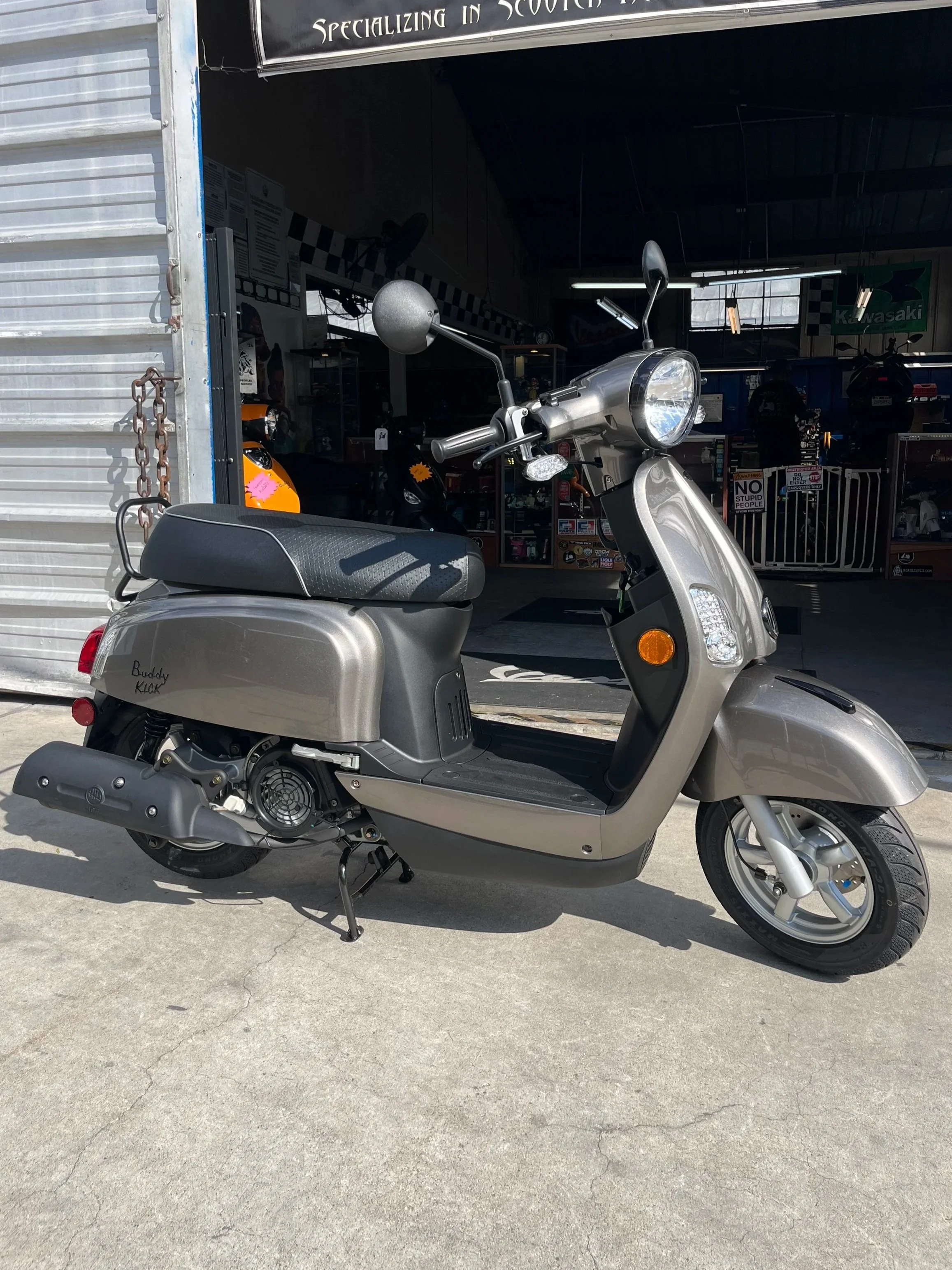 Silver scooter parked outside a scooter shop with an open garage door showing helmets and merchandise inside.