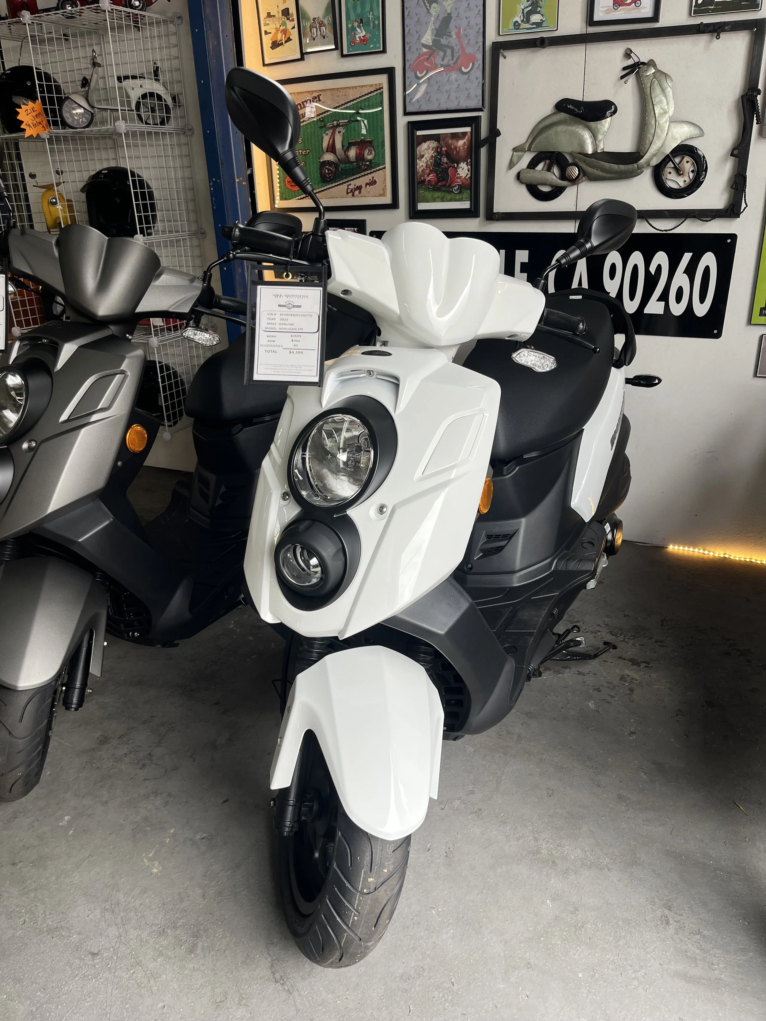 A white scooter with a black seat and handlebar, displayed inside a showroom next to other scooters, with framed motorcycle artwork on the wall behind.