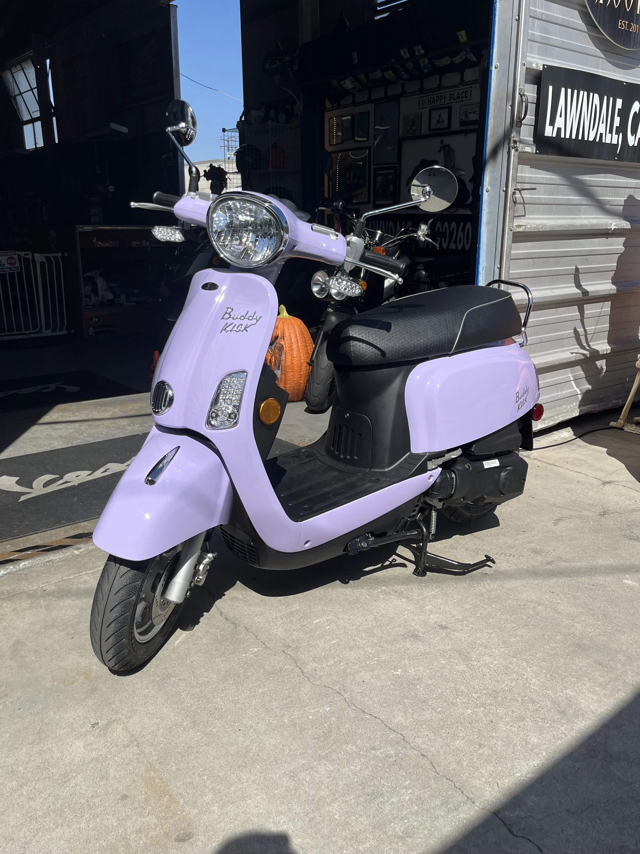 A light Lavender scooter with a black seat parked outside a store with a partially open door, visible inside are various framed pictures and decorations, with a pumpkin-shaped object nearby.