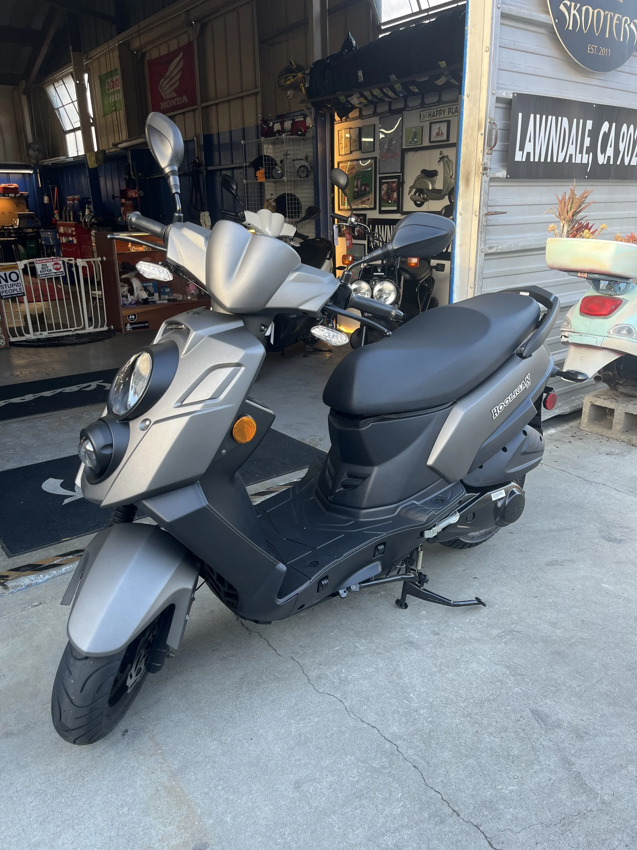 A silver and black scooter parked outside an indoor shop with various items and framed pictures on the wall. The shop has signs including one that says 'LAWNDale, CA 902' and another with 'SKOOTER' with 'EST. 2011.'
