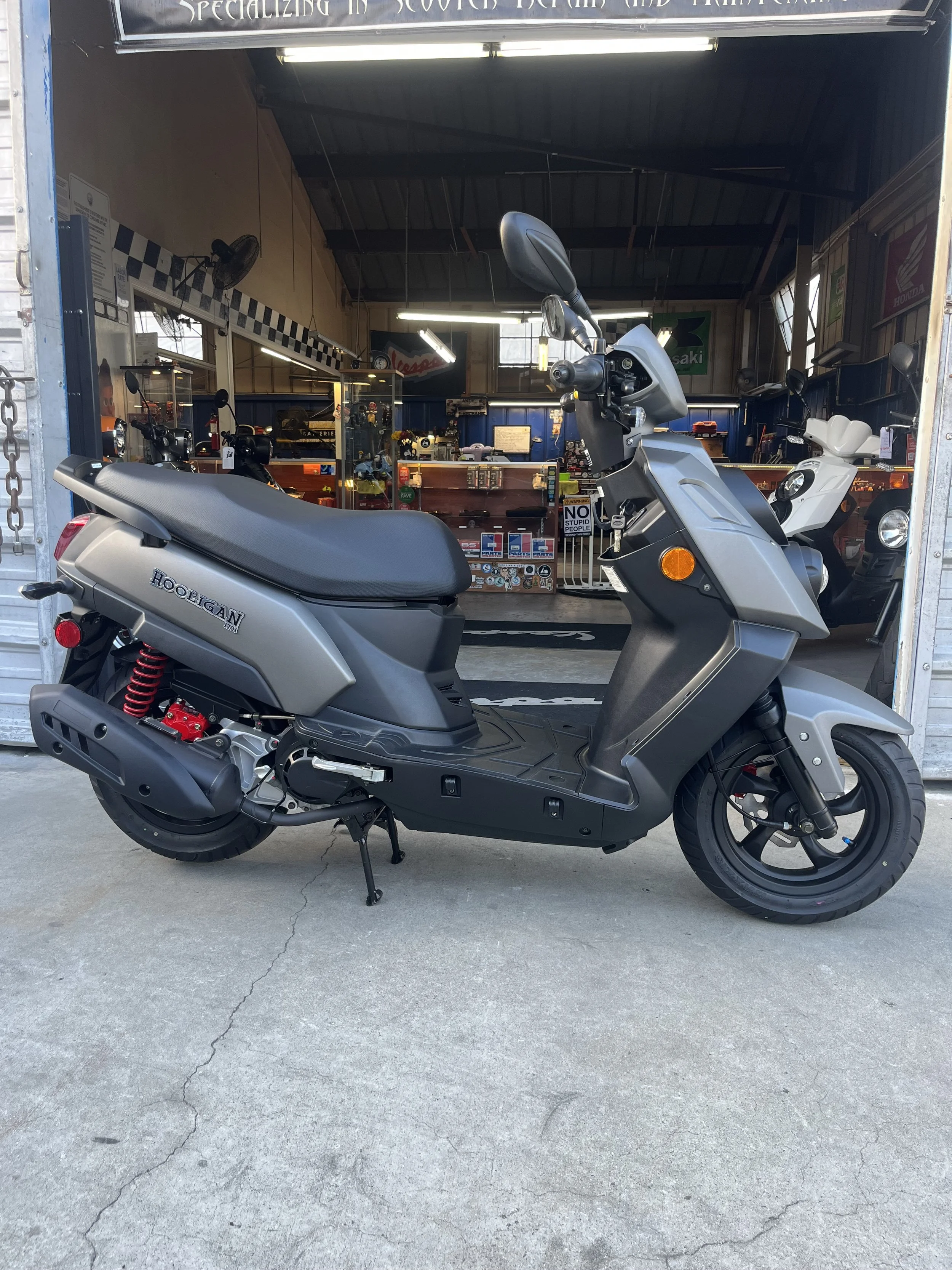 Silver motor scooter parked outside a motorcycle shop with bikes and accessories inside.