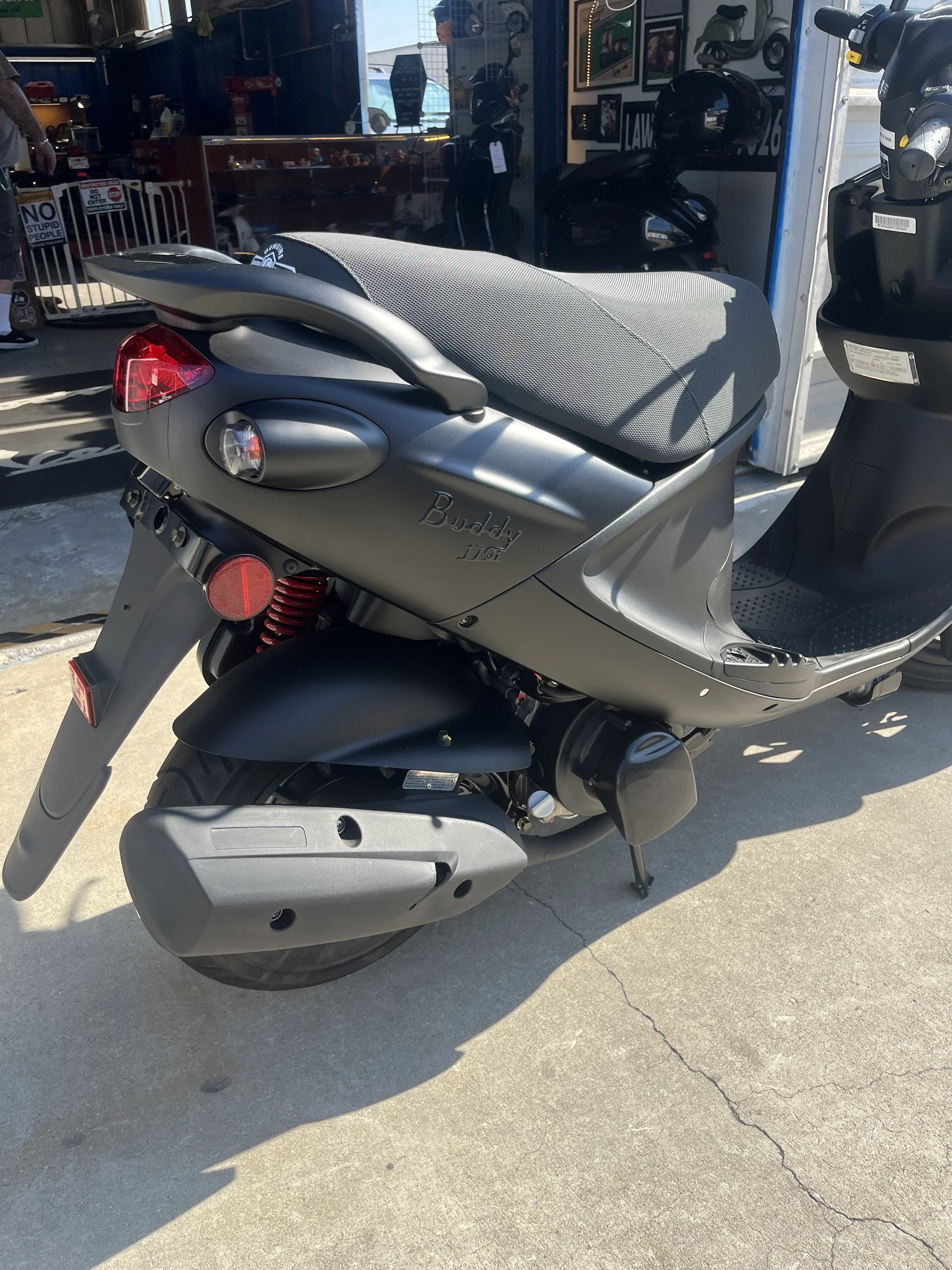 Gray Buddy scooter parked outside a shop with a glass front, showcasing motorcycle gear and framed pictures on the wall inside.