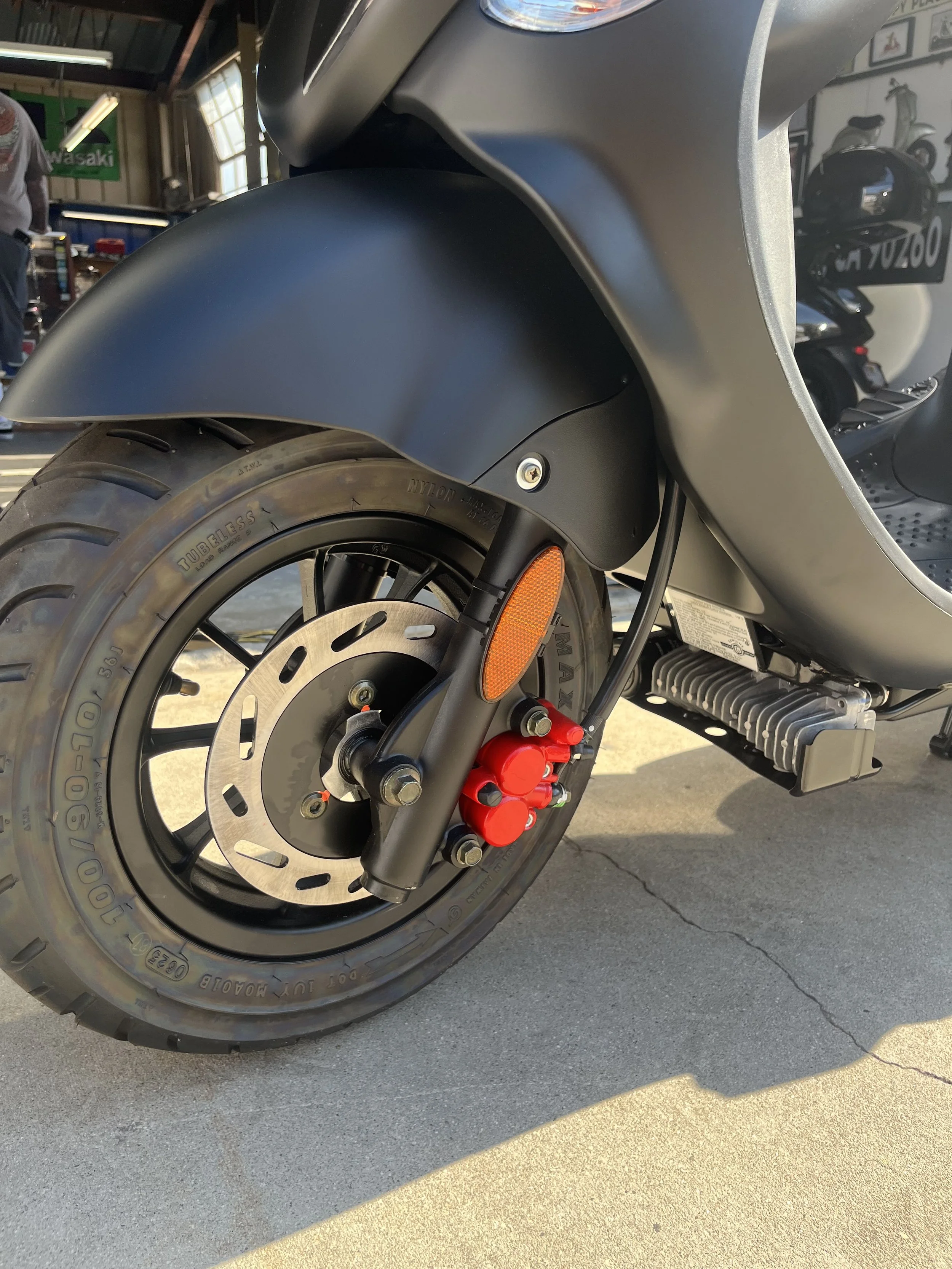 Close-up of the front wheel of a motorcycle, showing the tire, brake disc, red brake caliper, and front fender, with part of the engine visible in the background.