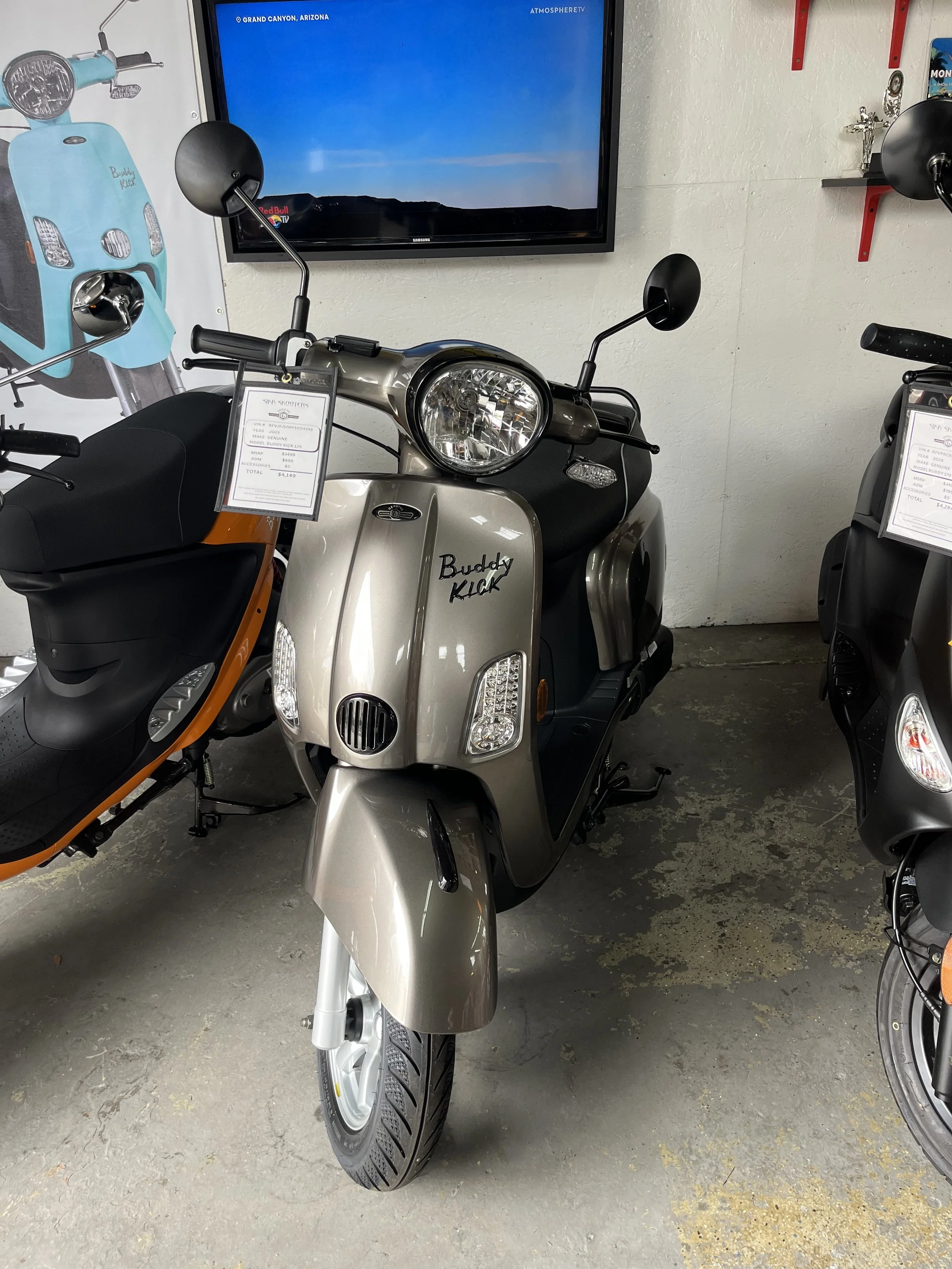 A silver and black scooter labeled 'Buddy Kick' on display in a showroom, with a wall-mounted TV behind it showing Grand Canyon, Arizona, and other scooters with price tags on either side.