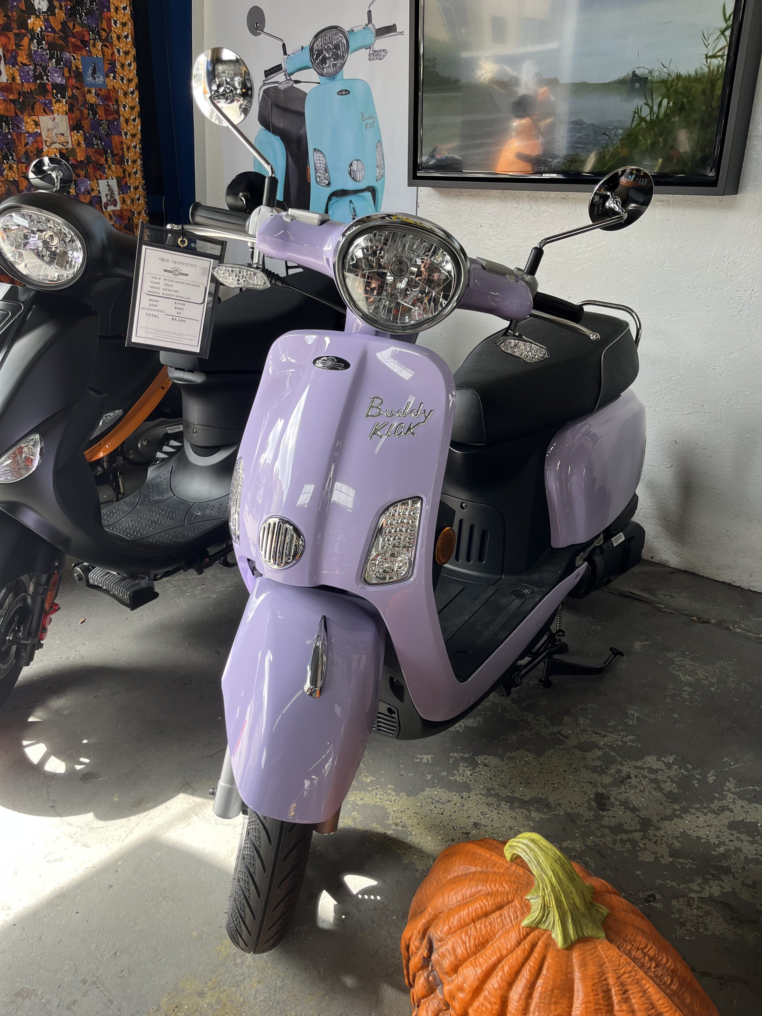 A purple Buddy Kick electric scooter on display indoors, with a decorative pumpkin on the ground in front of it, next to a black scooter, with a blue mural of a vintage scooter and a television in the background.