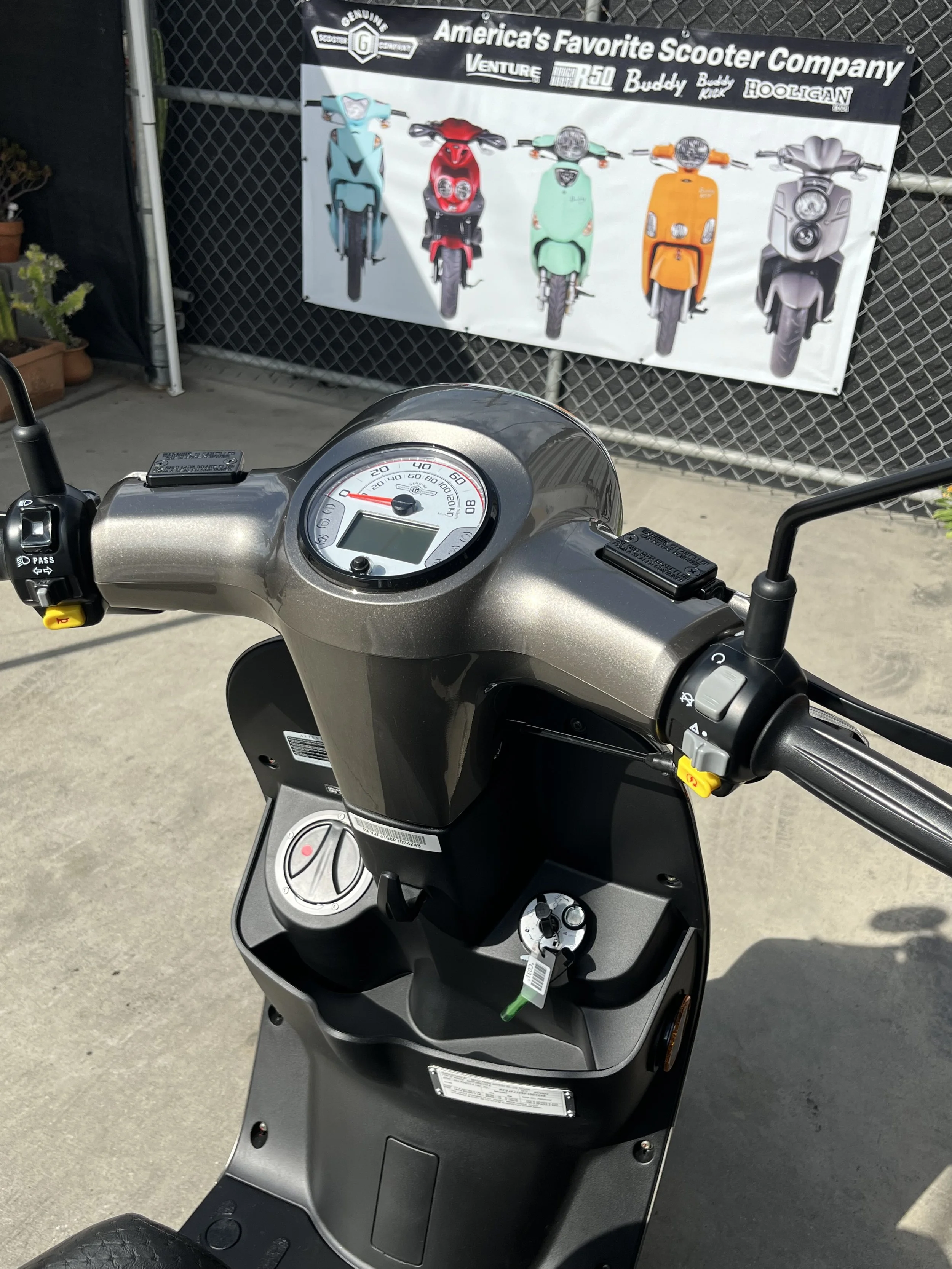 View of a black scooter's handlebar and dashboard with a key inserted, parked outside near a chain-link fence. Behind the scooter, a banner displays various colorful scooters and the text 'America's Favorite Scooter Company'.