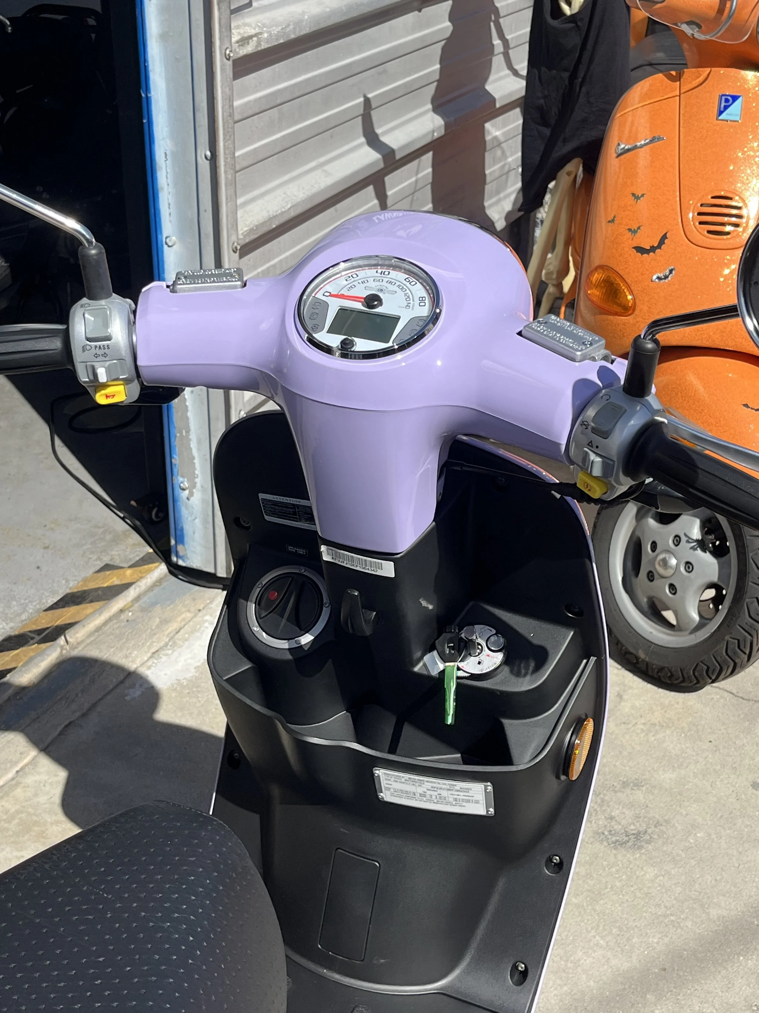 Close-up of a purple scooter's dashboard, showing a speedometer, ignition key, and handlebar controls, with an orange scooter and a person in the background.