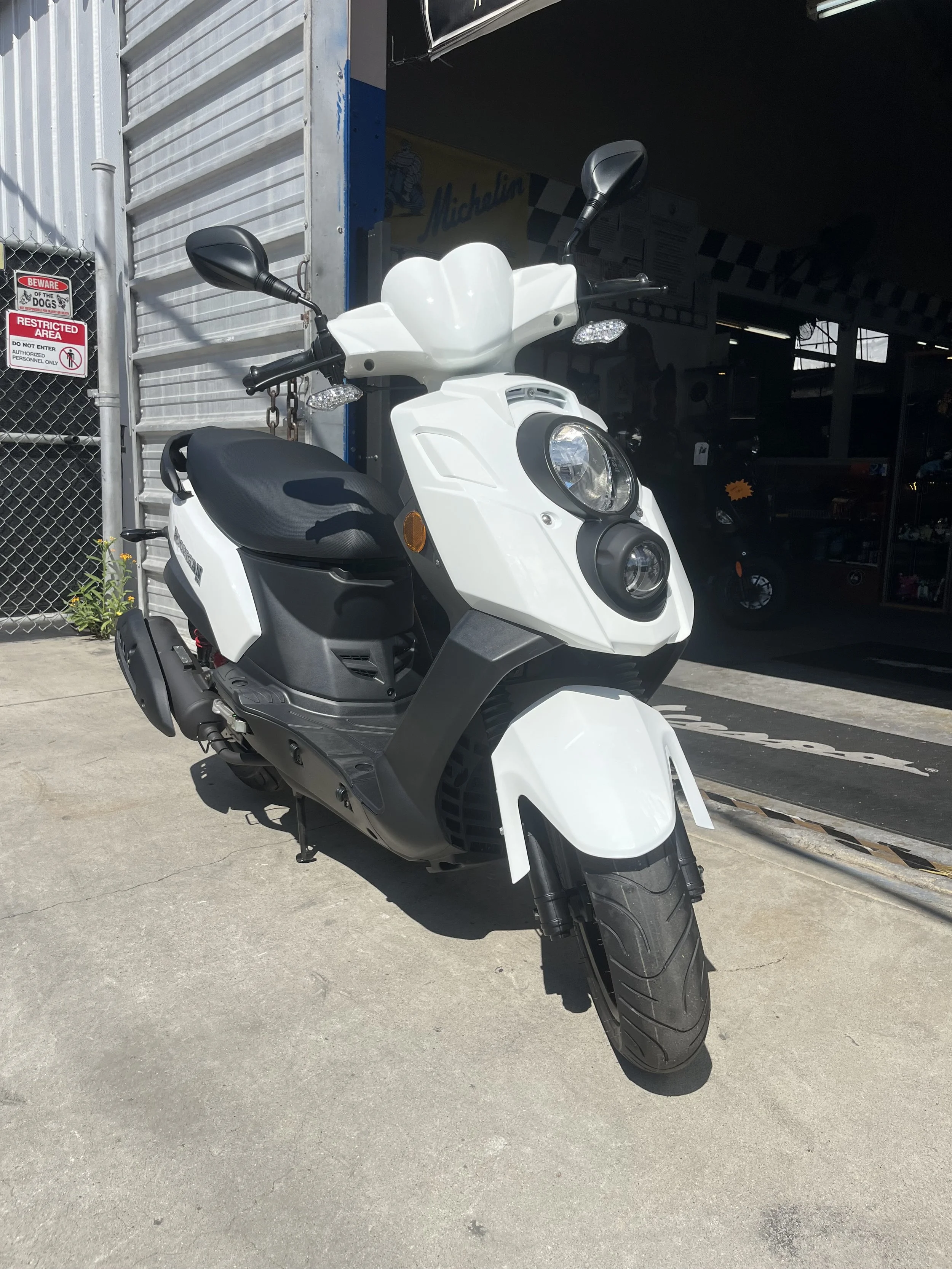 White electric scooter parked outside a building with a black door, dark interior, and a yellow 'Michelin' sign visible inside.