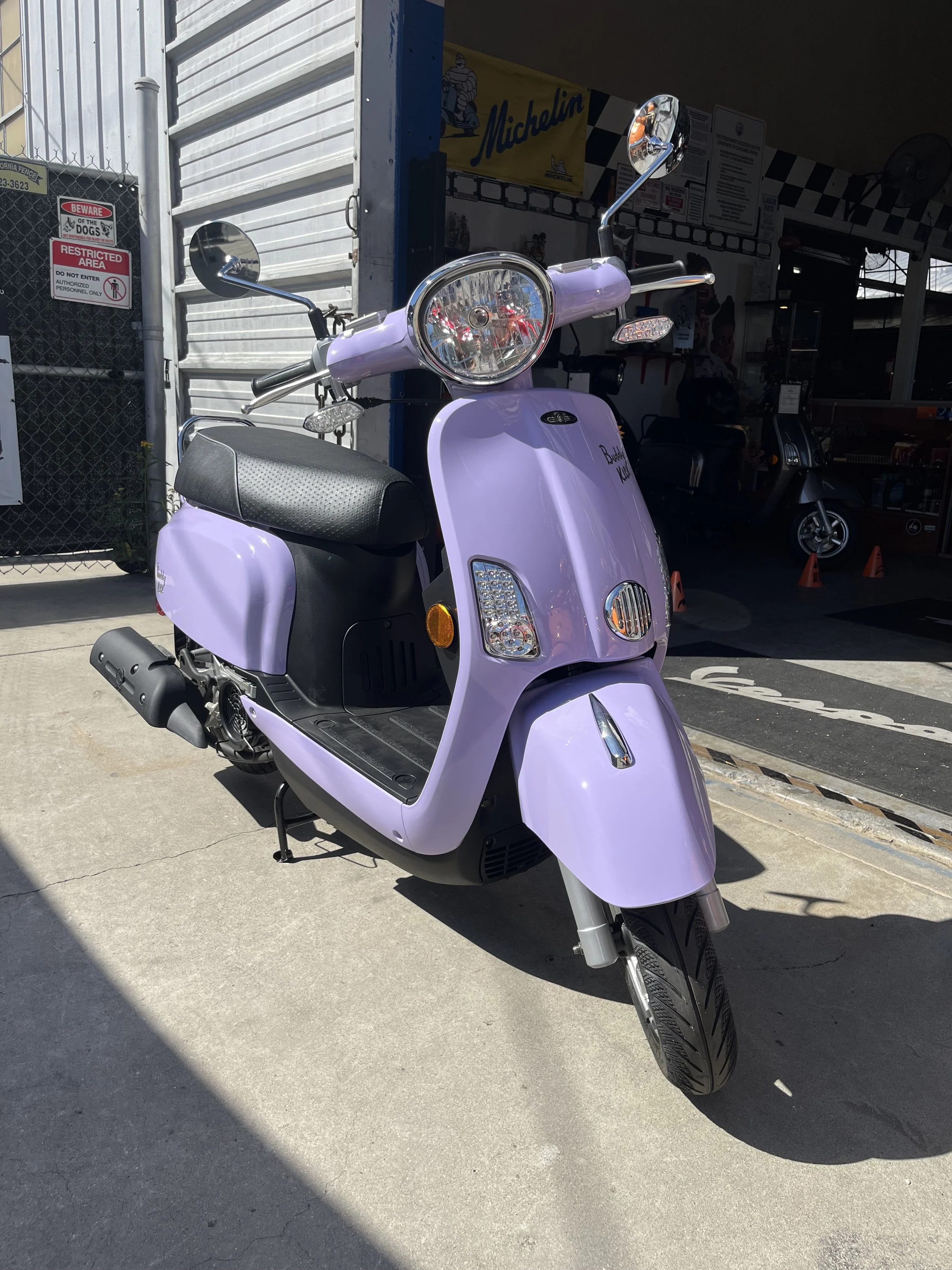 A lavender-colored scooter parked outside a building with a Michelin Sign in the background.