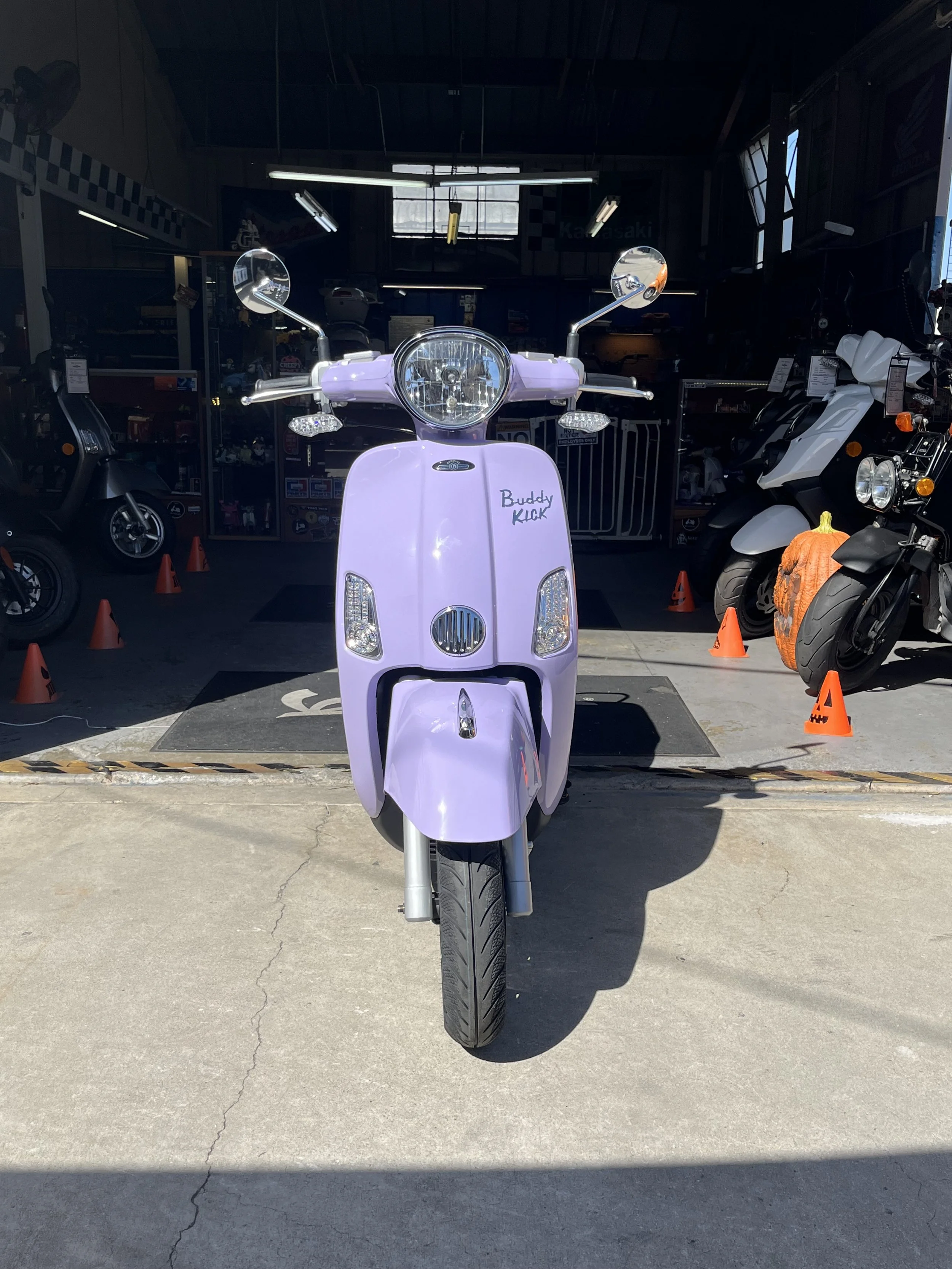 Front view of a lavender-colored scooter parked outside a motorcycle shop, with the words 'Buddy KICK' written on the front.