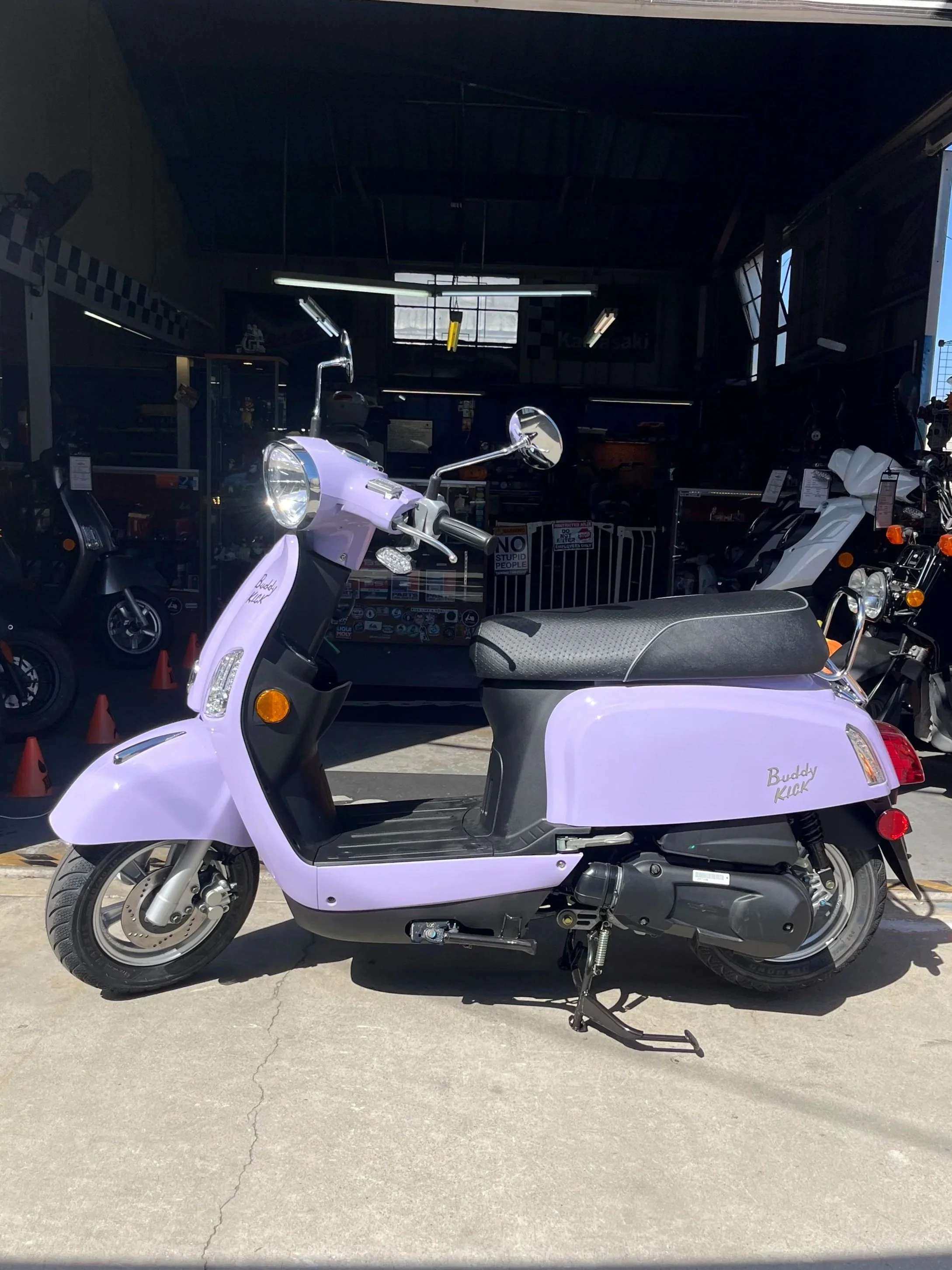 A light purple scooter with black seat and black details parked outside a shop with various scooters inside.