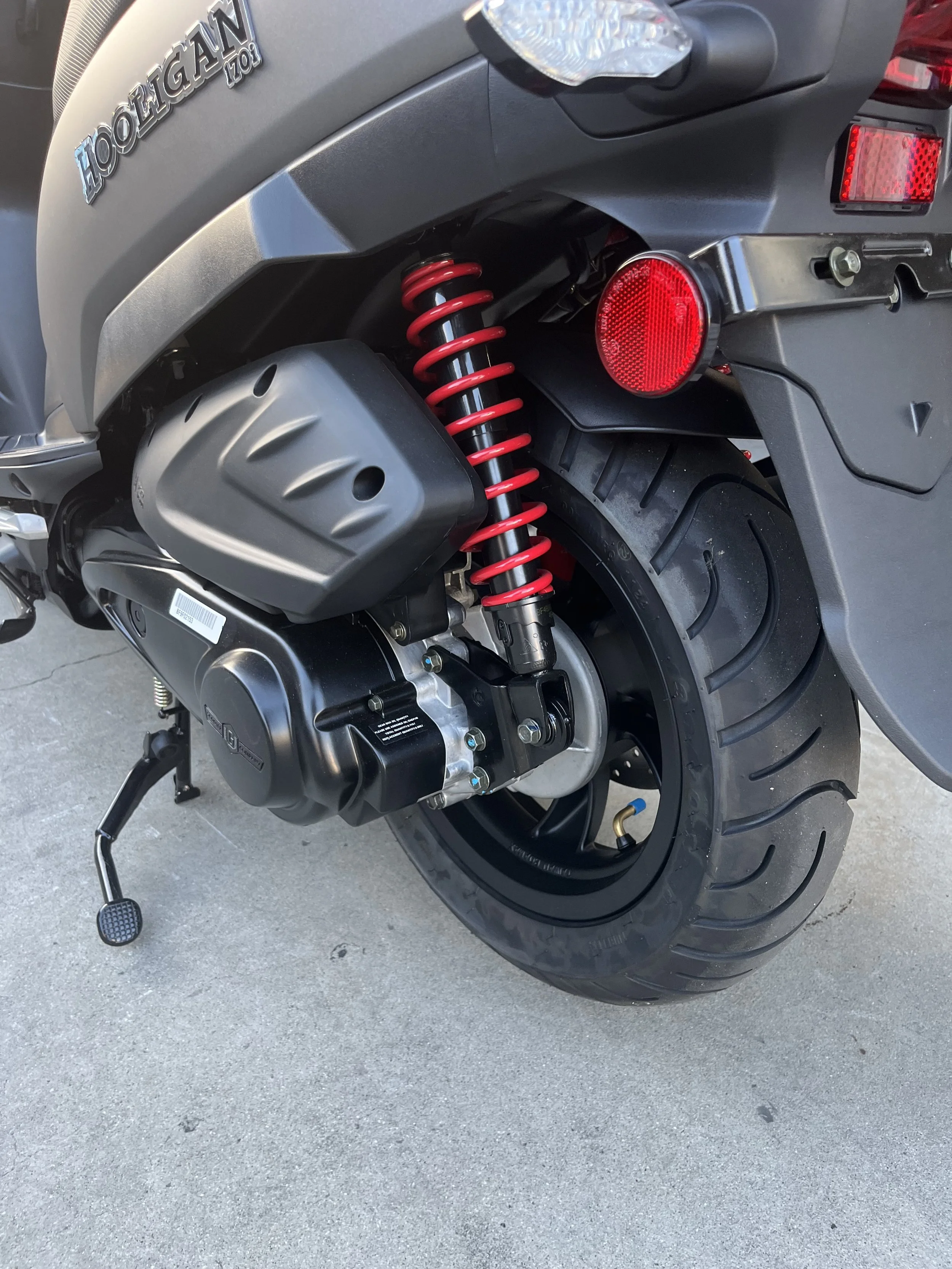Close-up of the rear wheel and suspension of a gray scooter with a red coil shock absorber, black engine cover, and a black tire.
