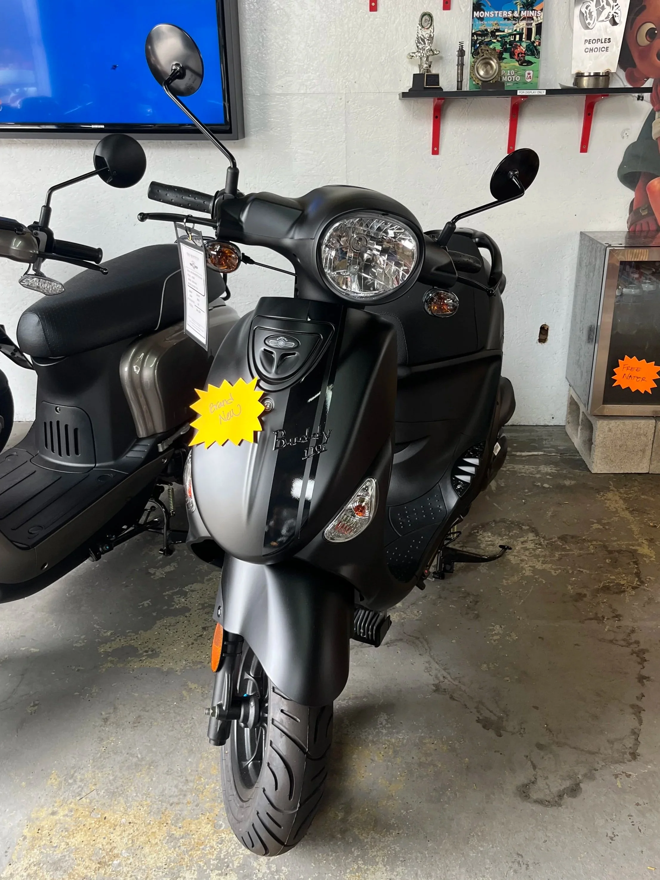 Black Buddy scooter with a yellow sale tag, displayed indoors near other scooters and accessories on shelves.