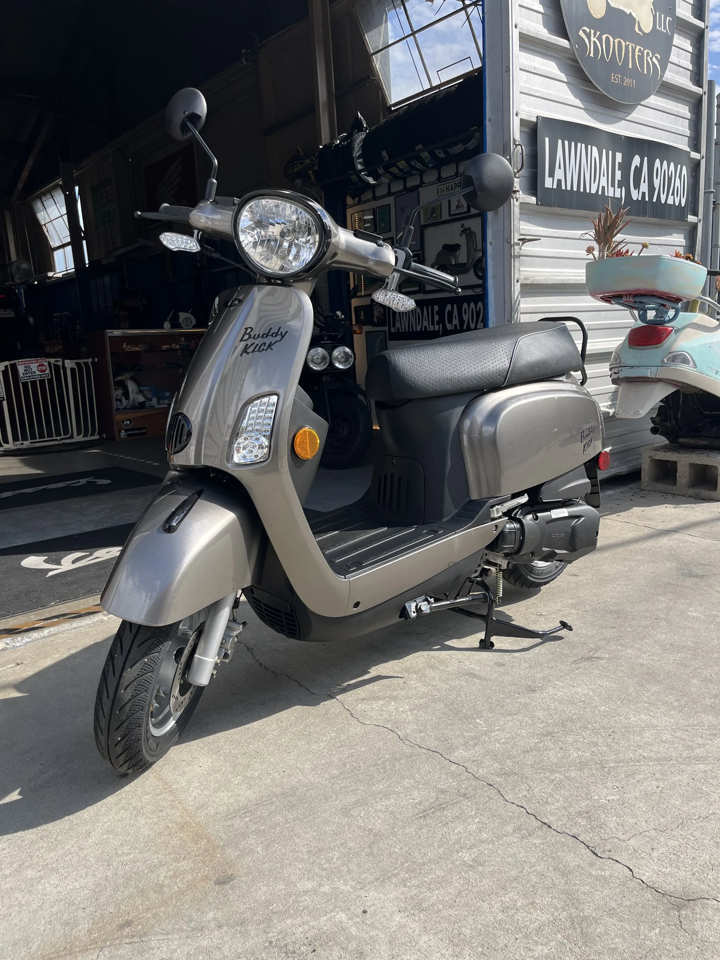 Glossy Titanium scooter with black seat parked outside a shop, with signs promoting Lawndale, California, and a sign reading 'Buddy Kick' on the scooter's front.