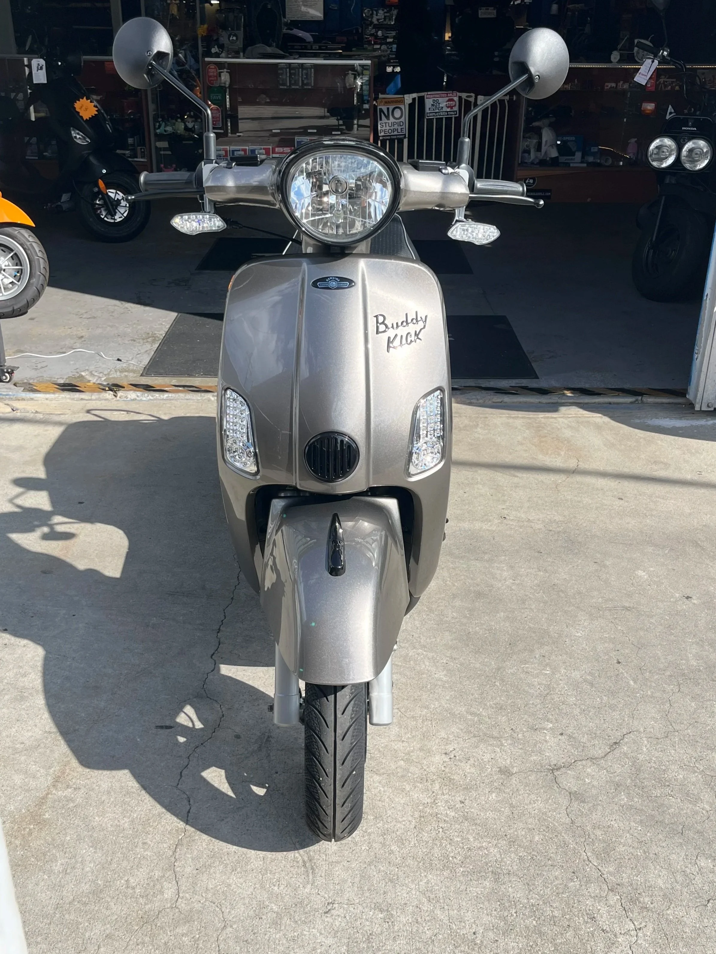 Front view of a silver scooter with the words "Buddy Kick" written on the front panel, parked outside a shop.