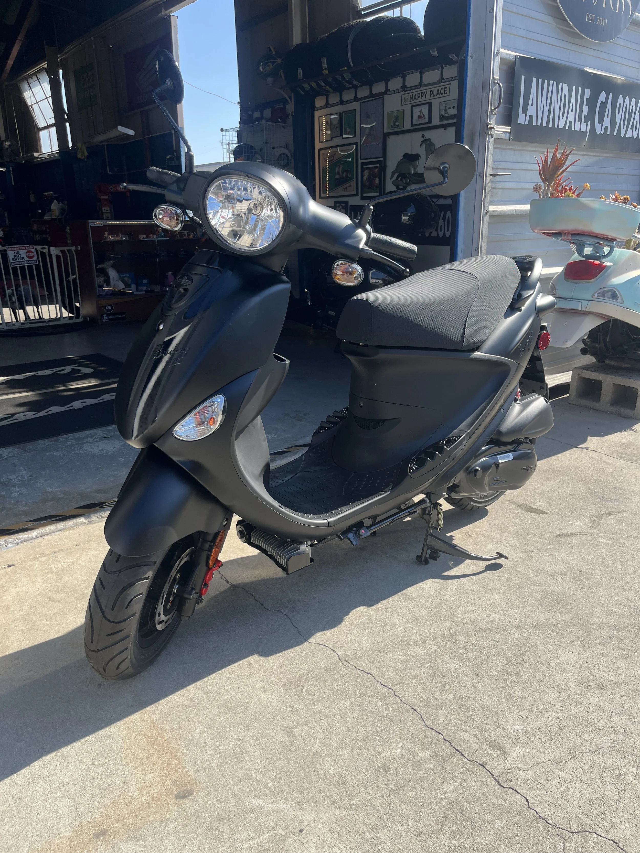 Black Buddy 170 scooter parked outside a shop with a sign reading 'Lawndale, CA' and a basket with plants on the back.
